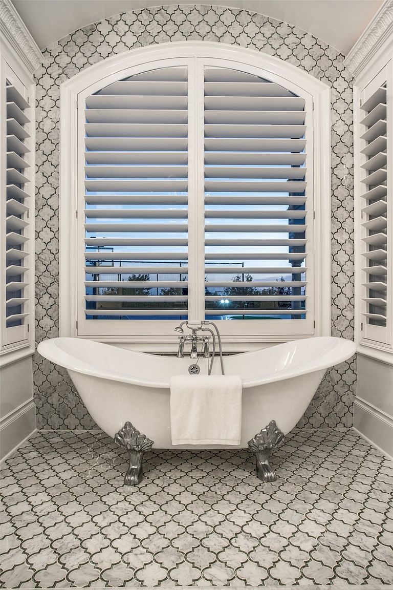 This elegant primary bathroom features a classic clawfoot soaking tub centered beneath a large, arched window with white plantation shutters. The walls and floor are adorned with a sophisticated, interlocking patterned tile, creating a cohesive and luxurious aesthetic. A polished metal faucet assembly is mounted on the tub, and a crisp white towel is draped over the side, emphasizing a spa-like atmosphere.