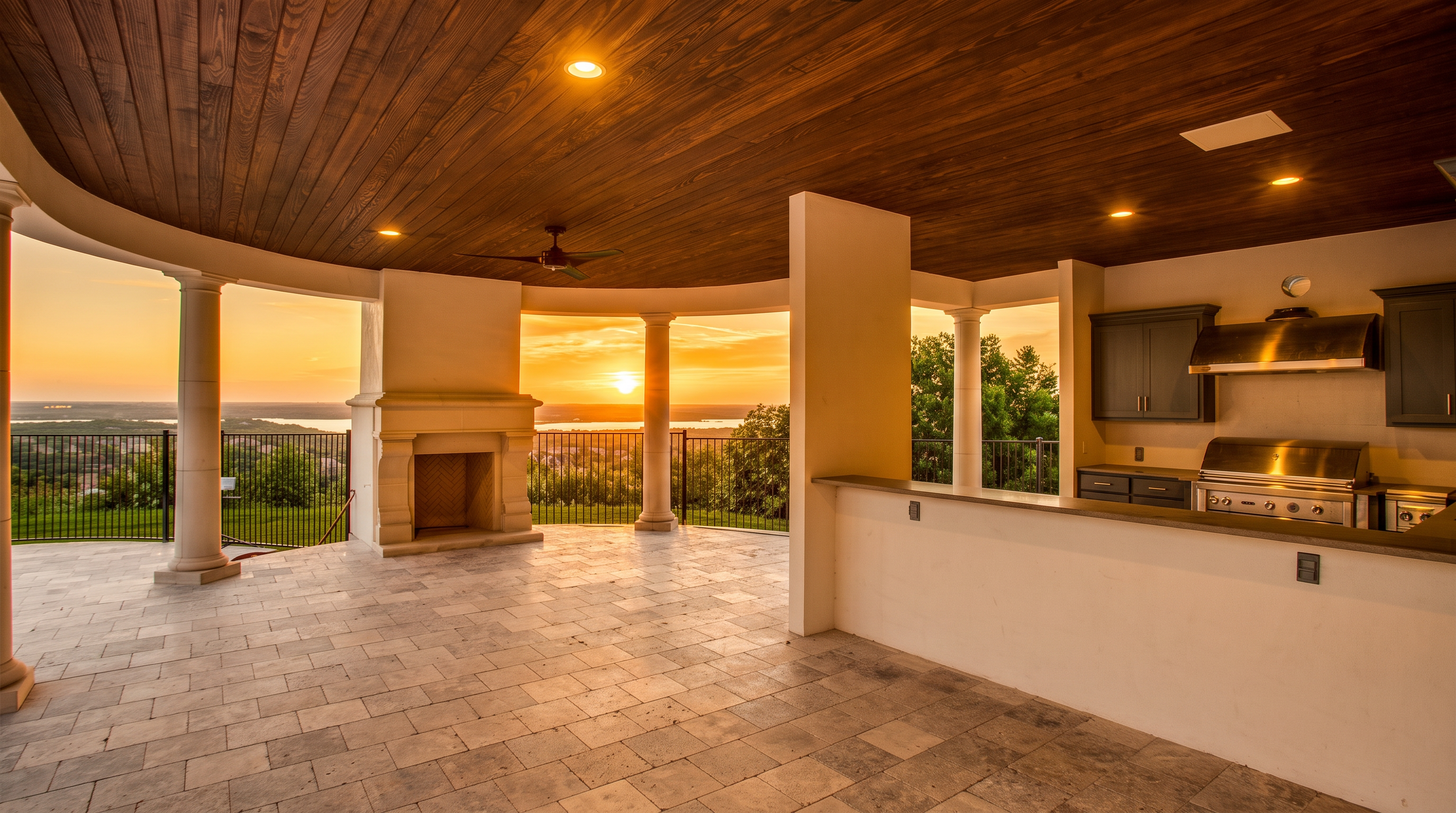 This expansive covered outdoor living space features a curved design with a rich wood-paneled ceiling and stone tile flooring. The area is equipped with a built-in outdoor kitchen, a stone fireplace, and classic columns that frame a panoramic view of the landscape and water below. The open-air layout creates a seamless transition between the indoor-style amenities and the scenic outdoor environment.