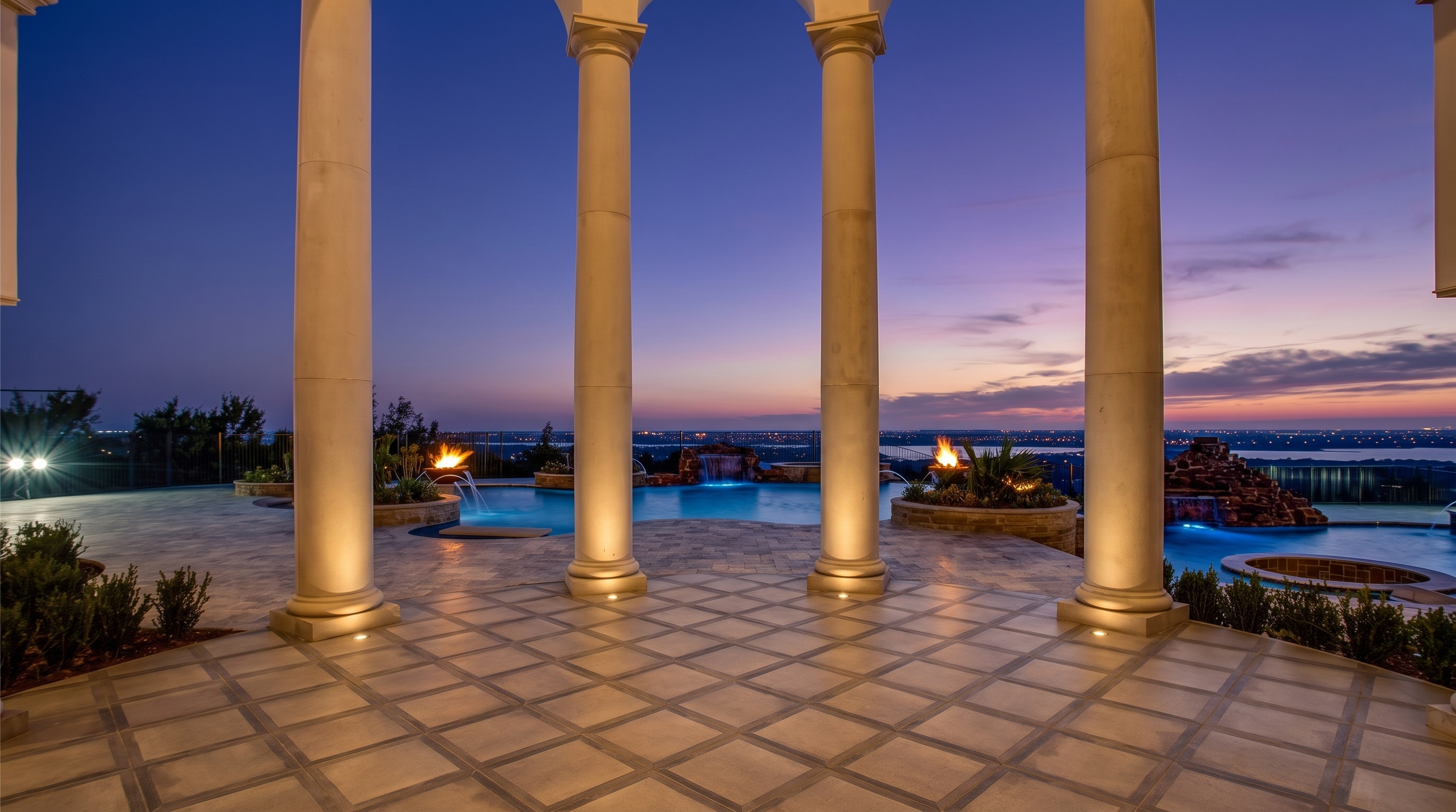 This luxurious covered patio features a classic colonnade with elegant white pillars framing a stunning panoramic view of a distant city skyline at dusk. The foreground showcases a tiled terrace leading to a multi-level swimming pool area complete with integrated water features and fire bowls. The scene captures a sophisticated outdoor living space designed for high-end entertaining with a dramatic sunset backdrop.