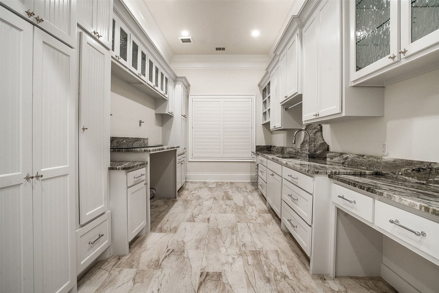 This bright and spacious laundry room features extensive white cabinetry, a deep sink with a gooseneck faucet, and patterned stone countertops. The room is finished with light-toned, large-format tile flooring and a central window with shutters that allows for natural light. The symmetrical layout provides ample counter space and storage, creating a clean and functional utility area.