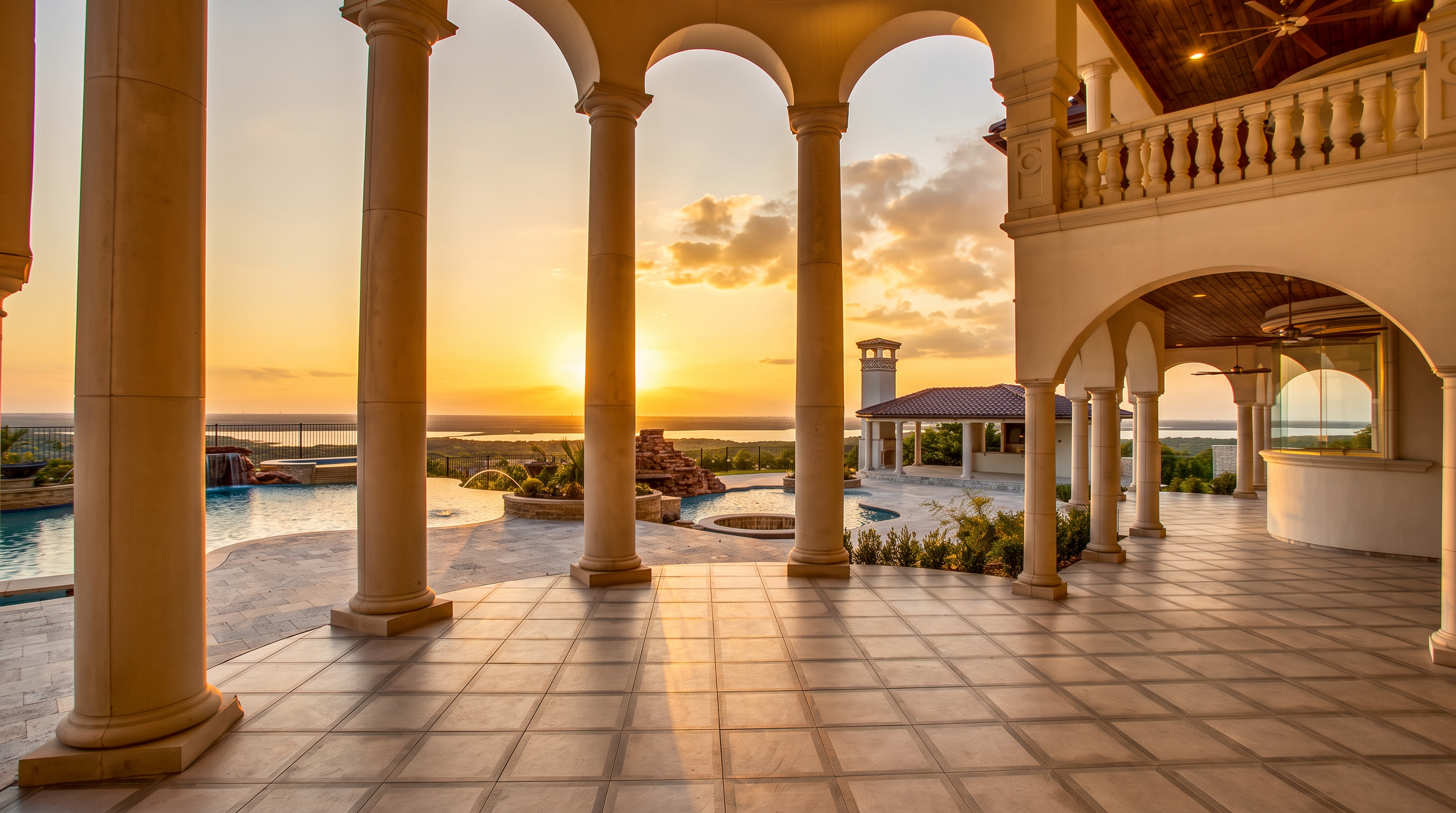 This expansive covered patio features a series of grand, classical columns that frame a breathtaking sunset view over a distant lake. The space is paved with large, grid-patterned tiles and includes a multi-level swimming pool with a rock waterfall feature in the background. The architectural design incorporates a high, wood-paneled ceiling and a stone-clad chimney structure, creating a luxurious and serene outdoor living environment.