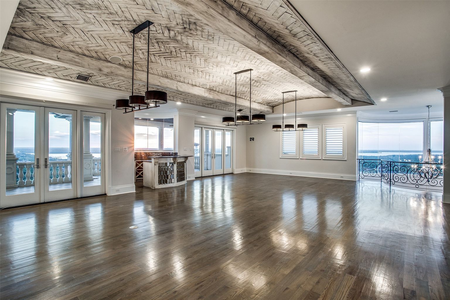 This expansive living area features a striking vaulted ceiling with a herringbone-patterned wood finish and exposed beams. The room is anchored by polished hardwood floors and illuminated by three modern, multi-light pendant chandeliers. Large glass doors and windows provide panoramic views of the surrounding landscape, creating a bright and airy atmosphere with a sophisticated, open-concept layout.