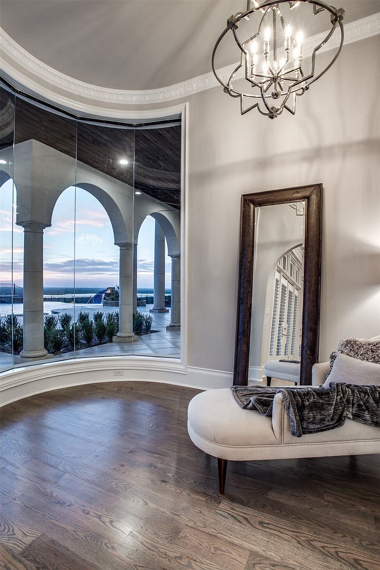 This elegant sunroom features a curved wall of floor-to-ceiling glass that provides an expansive view of a covered patio and a pool area at dusk. The space is furnished with a plush, light-colored chaise lounge topped with a dark throw blanket, positioned near a large, dark-framed floor mirror. A sophisticated, multi-tiered metal chandelier hangs from the ceiling, casting a warm glow over the dark wood flooring.