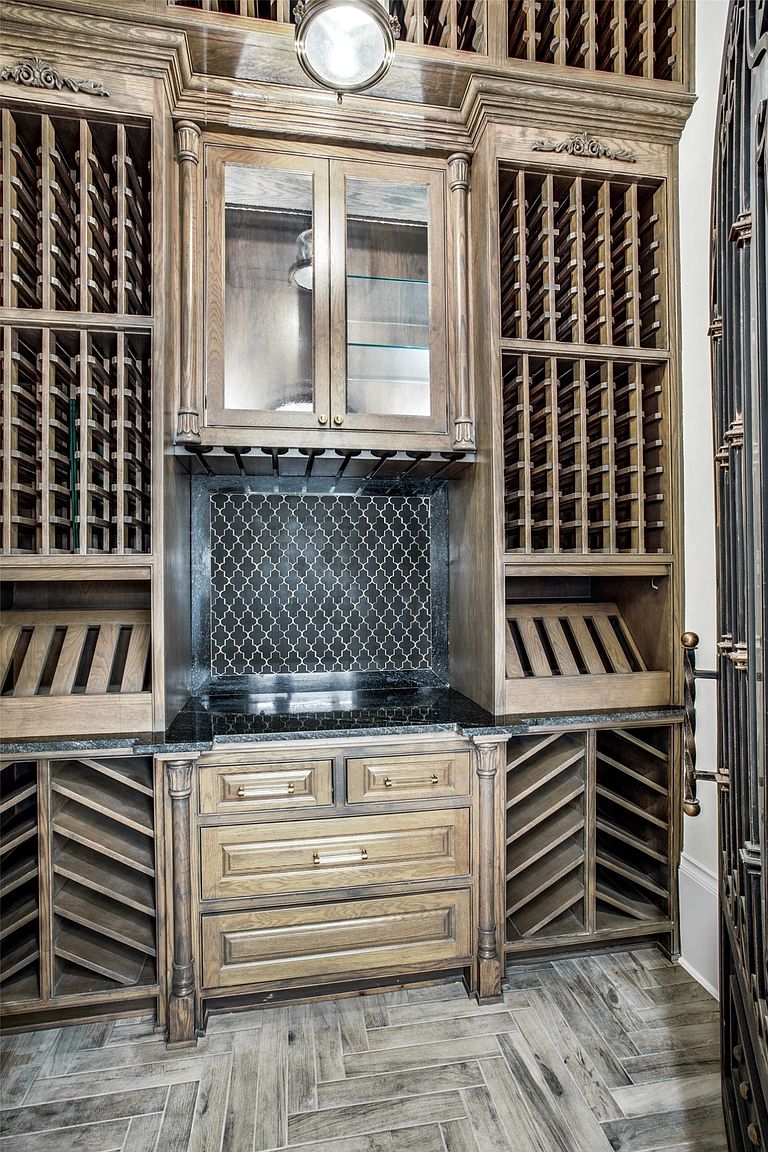 This sophisticated wine cellar features extensive custom wood cabinetry with dedicated bottle storage racks, display shelves, and a central glass-fronted cabinet. A dark, patterned tile backsplash sits above a stone countertop, providing a refined contrast to the warm-toned wood. The space is finished with a herringbone-patterned floor and an ornate metal gate, creating an elegant and functional area for wine collection storage.