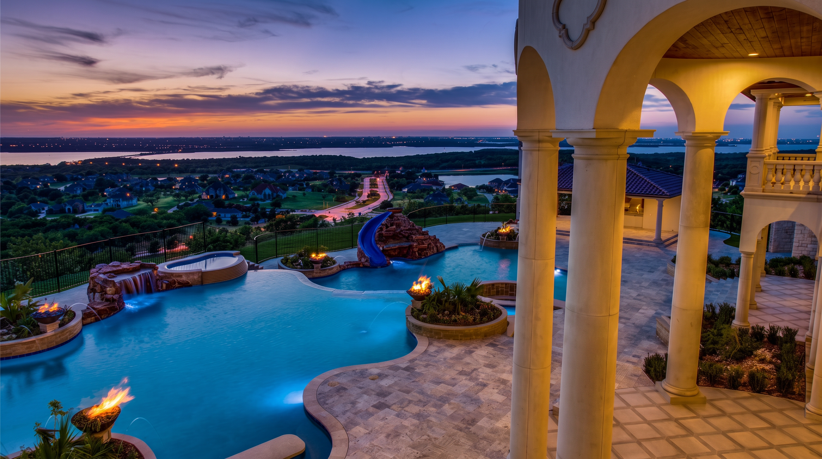 This stunning elevated view captures a luxurious infinity-edge pool and spa complex at twilight, overlooking a sprawling suburban landscape and distant lake. The scene features a multi-level stone patio, a blue water slide, and integrated fire features that cast a warm glow against the cool evening sky. The perspective is framed by elegant, light-colored stone columns of a covered loggia, emphasizing the high-end architectural design and expansive outdoor living space.