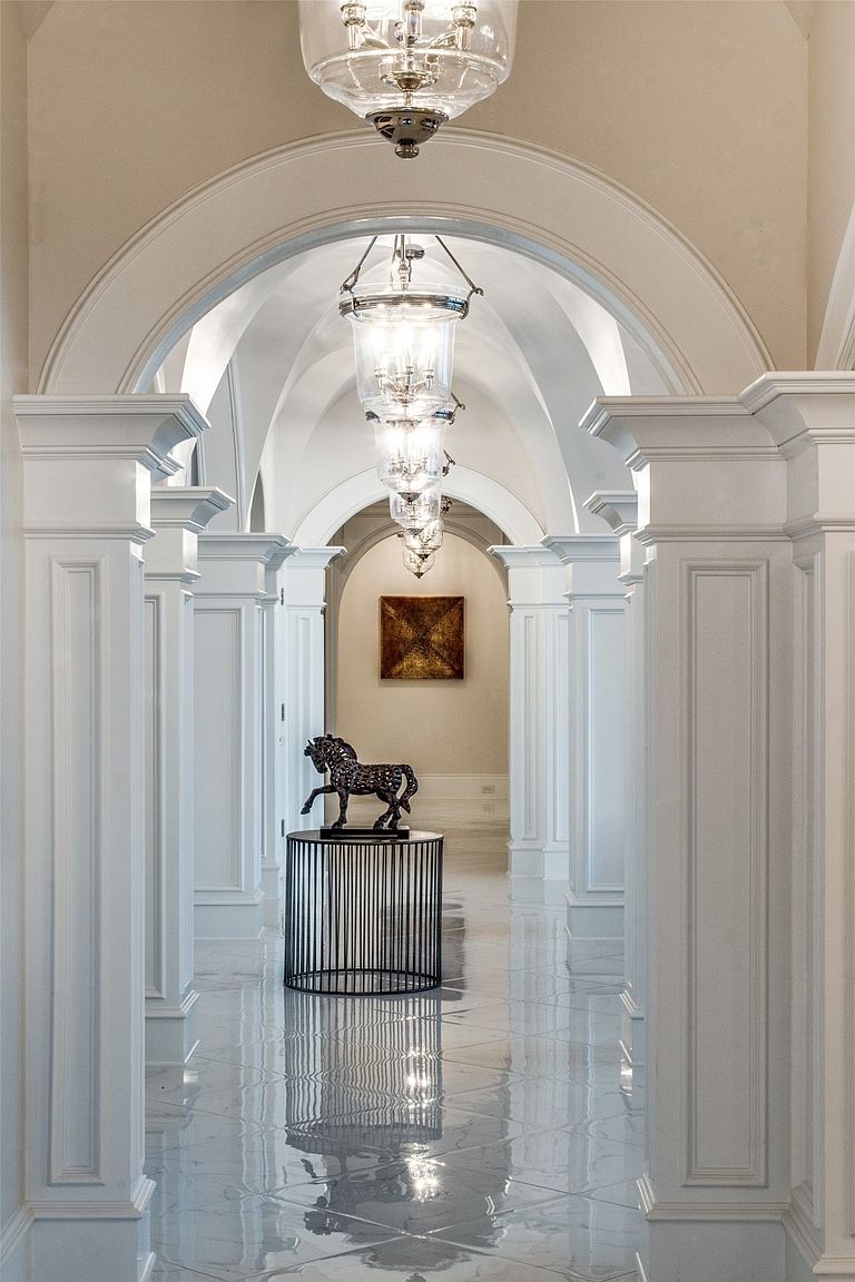 This elegant, long hallway features a series of arched architectural openings that create a dramatic, rhythmic perspective. The space is illuminated by a sequence of hanging glass lanterns and finished with polished, reflective light-toned flooring. A dark, circular pedestal table holding a horse sculpture serves as a focal point, while a piece of framed art hangs on the far wall, enhancing the sophisticated and symmetrical design.