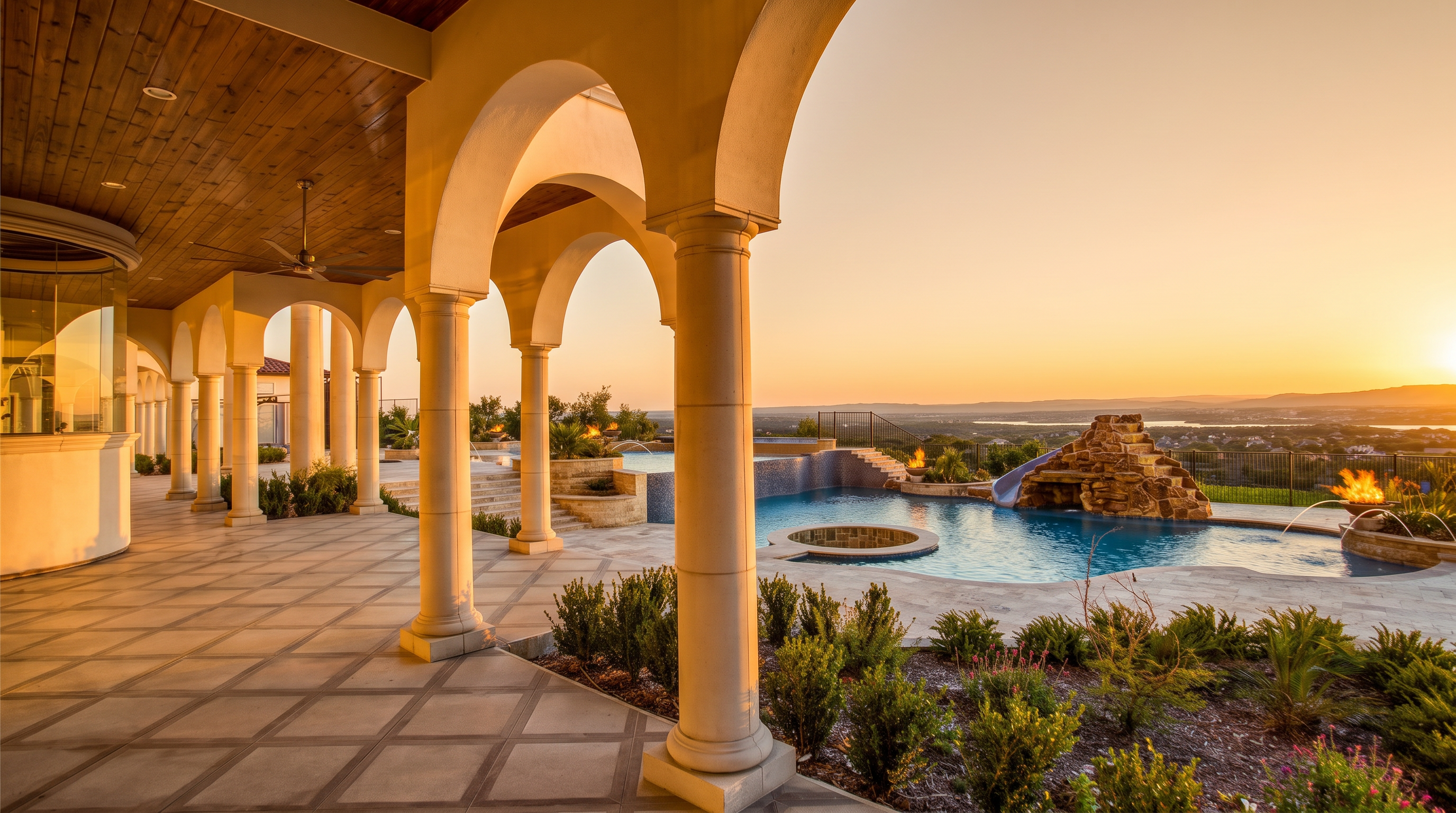 This expansive covered patio features a series of elegant arched columns and a warm-toned wood-paneled ceiling, creating a sophisticated outdoor living space. The patio overlooks a luxurious infinity-edge pool with a rock waterfall feature and a built-in spa, all set against a breathtaking panoramic sunset view of the horizon. The scene is captured from a low-angle perspective, emphasizing the architectural grandeur of the home and the seamless transition between the indoor-outdoor living areas.