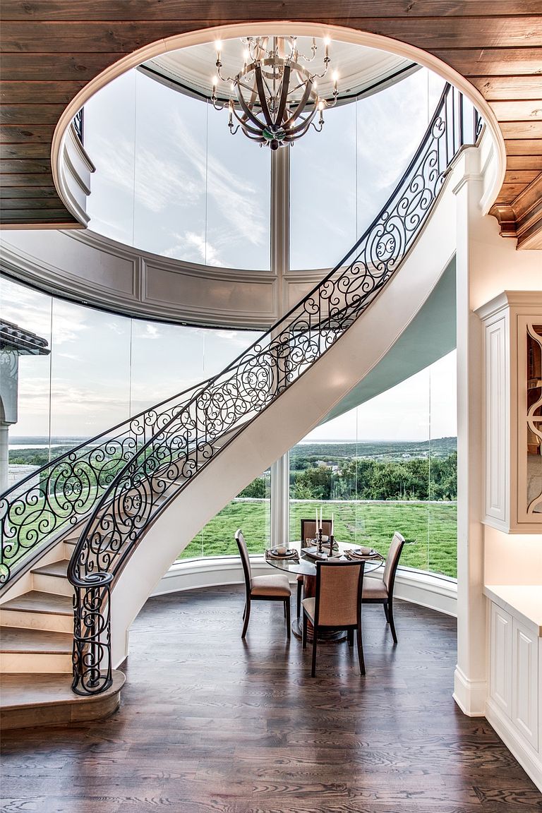 This elegant dining area is defined by a dramatic curved staircase with intricate wrought-iron railings that sweeps upward beneath a circular architectural cutout. The space features floor-to-ceiling glass walls that provide expansive, panoramic views of the surrounding landscape and sky. A round dining table with three upholstered chairs sits centrally on dark wood flooring, illuminated by a sophisticated chandelier hanging from the upper level.