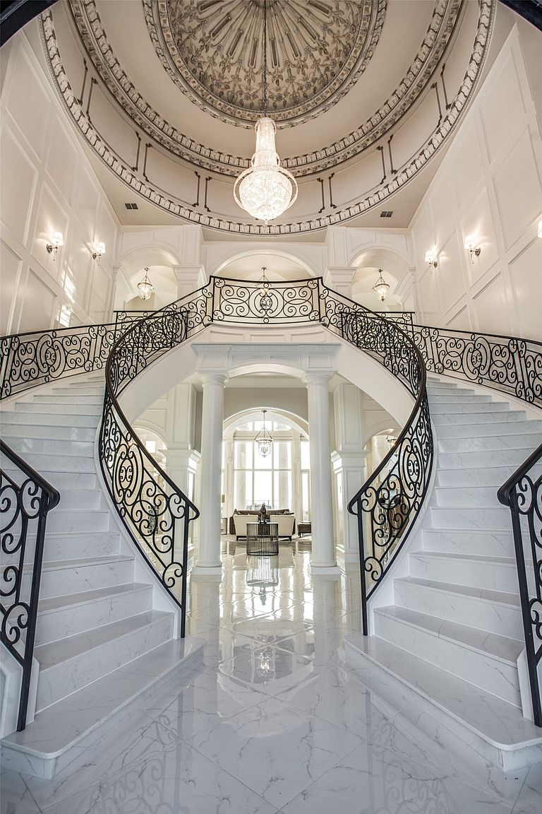 This grand foyer features a dramatic double staircase with intricate wrought-iron railings and light-colored stone steps. Above, a large crystal chandelier hangs from a detailed, circular domed ceiling, creating a sense of opulence and scale. The perspective looks directly through the center of the stairs toward a bright, open living area in the background, emphasizing the home's expansive and symmetrical architectural design.