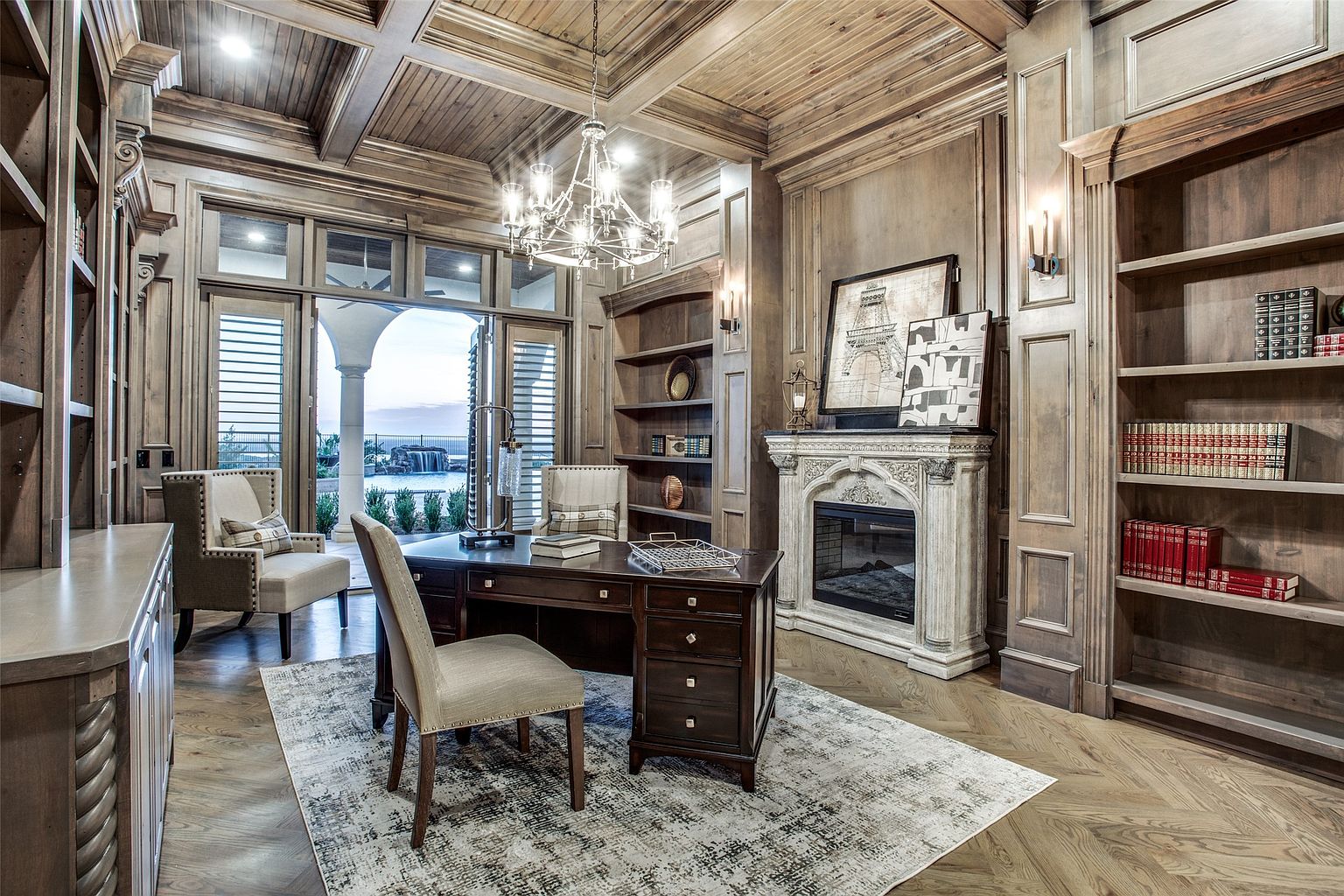 This sophisticated home office features extensive wood paneling, coffered ceilings, and built-in bookshelves that create a warm, scholarly atmosphere. A central dark wood desk sits atop a distressed area rug, complemented by a stone fireplace surround and a pair of elegant armchairs. Large glass doors open to a view of a backyard pool, blending traditional interior craftsmanship with a seamless transition to outdoor living.