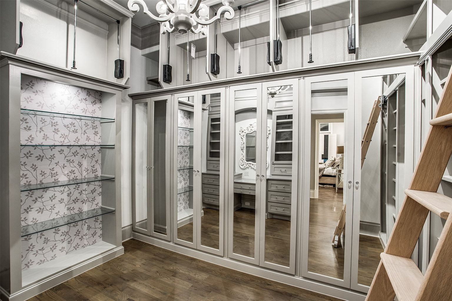 This luxurious walk-in closet features extensive custom cabinetry with mirrored doors and integrated lighting, creating a bright and sophisticated atmosphere. A rolling wooden ladder provides access to the upper storage sections, while a dedicated alcove with glass shelving and patterned wallpaper adds a decorative touch. The space is finished with rich wood flooring and a central chandelier, offering a high-end, organized aesthetic.