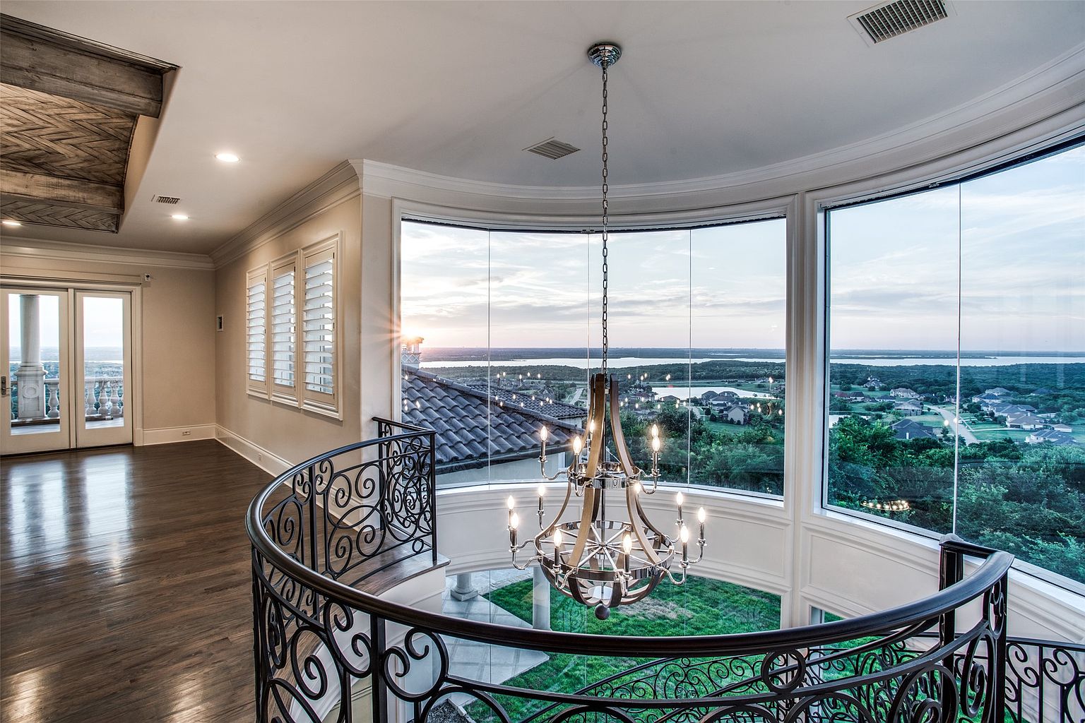 This elegant landing area features a curved wrought-iron railing overlooking a lower level, centered around a grand multi-tiered chandelier. Large, curved windows provide a panoramic view of the surrounding landscape and sunset, creating a bright and airy atmosphere. The space is finished with dark wood flooring and neutral walls, emphasizing a sophisticated and open architectural design.