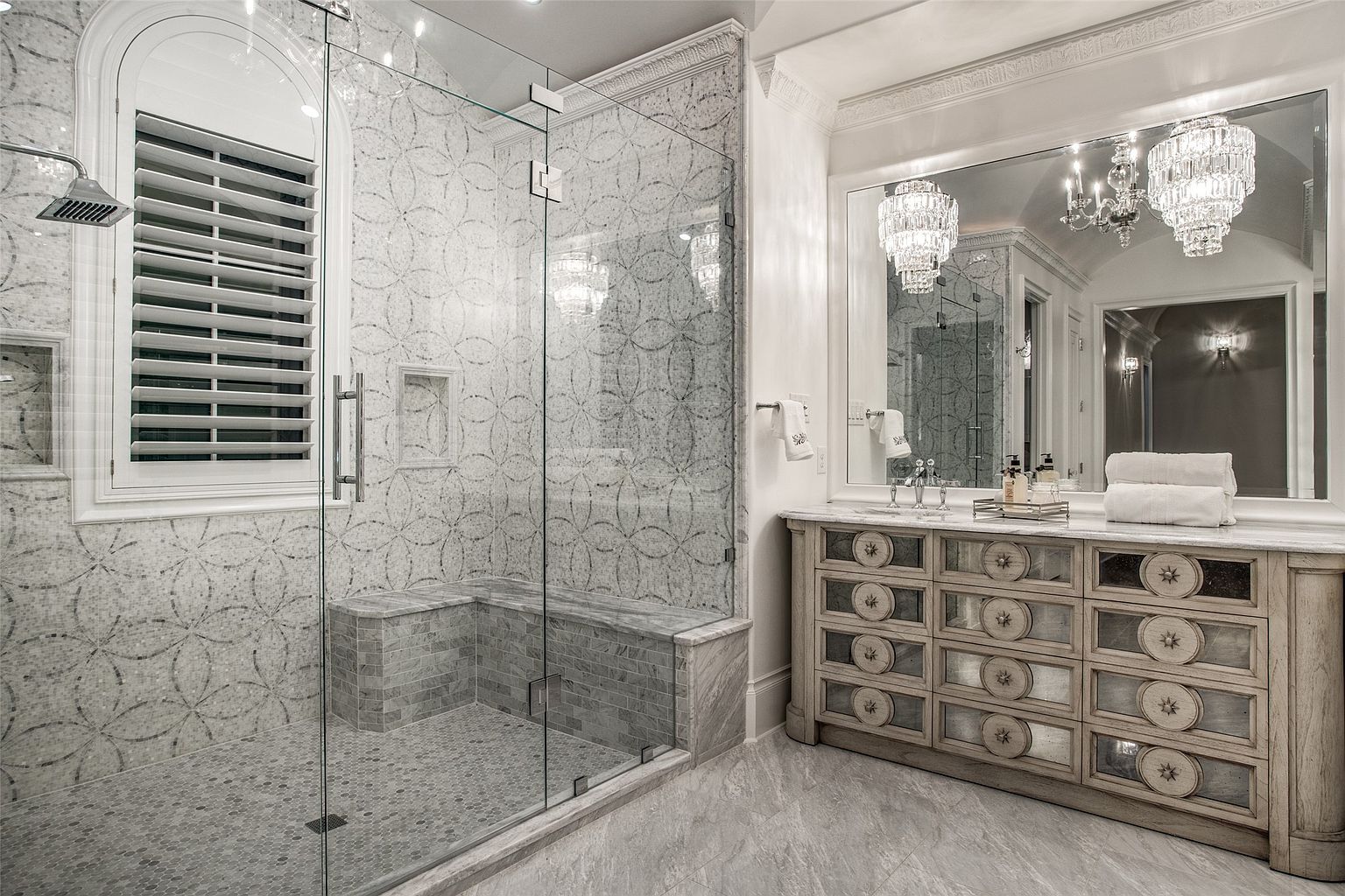 This luxurious primary bathroom features a spacious walk-in shower with intricate patterned tile walls and a built-in stone bench. A large vanity with decorative mirrored cabinet doors and a stone countertop sits beneath a wide mirror, complemented by elegant crystal chandeliers. The room is finished with light-toned stone flooring and a window with plantation shutters, creating a bright and sophisticated atmosphere.