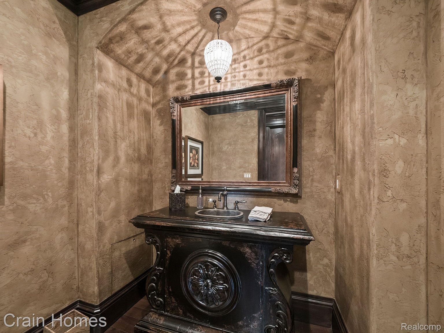 This is a powder room featuring a dark, ornate vanity with a vessel sink and a decorative mirror. The walls are textured with a brown, stucco-like finish, and a unique light fixture hangs from the ceiling, casting interesting shadows. The overall style is luxurious and somewhat rustic, creating a unique and memorable space.