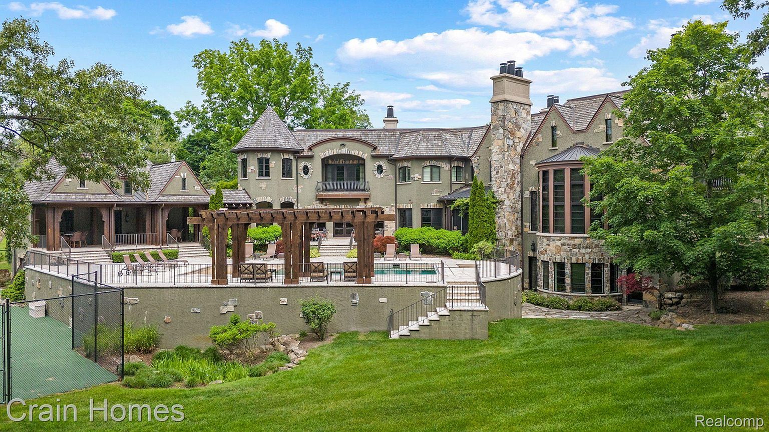 This is a rear view of a large, luxurious home featuring a stone and stucco exterior with multiple gables and a turret. The property includes a pool area with a pergola, a tennis court, and a well-manicured lawn. The overall impression is one of grandeur and privacy.