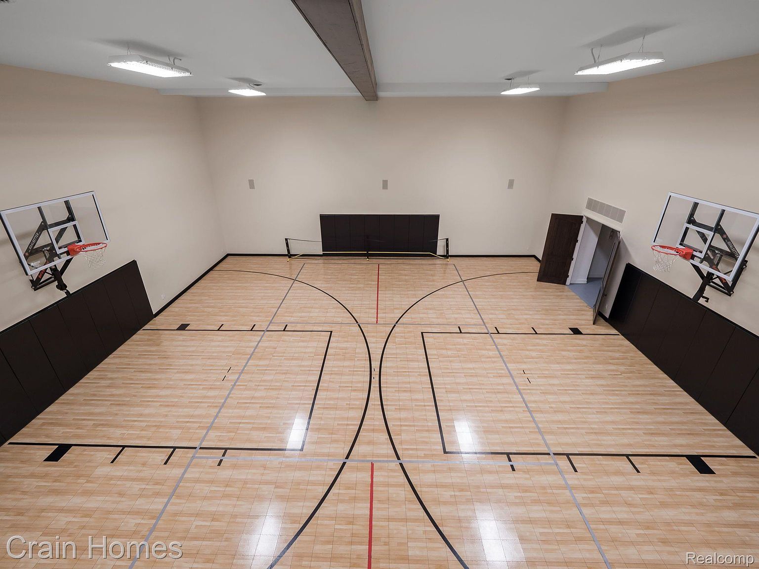 This is an indoor basketball court with hardwood flooring and wall-mounted basketball hoops on either side. The court features standard basketball court markings, including a center line, free throw lanes, and three-point arcs. The walls are painted in a neutral tone, and the space is well-lit with overhead lighting, creating a professional and inviting atmosphere for athletic activities.