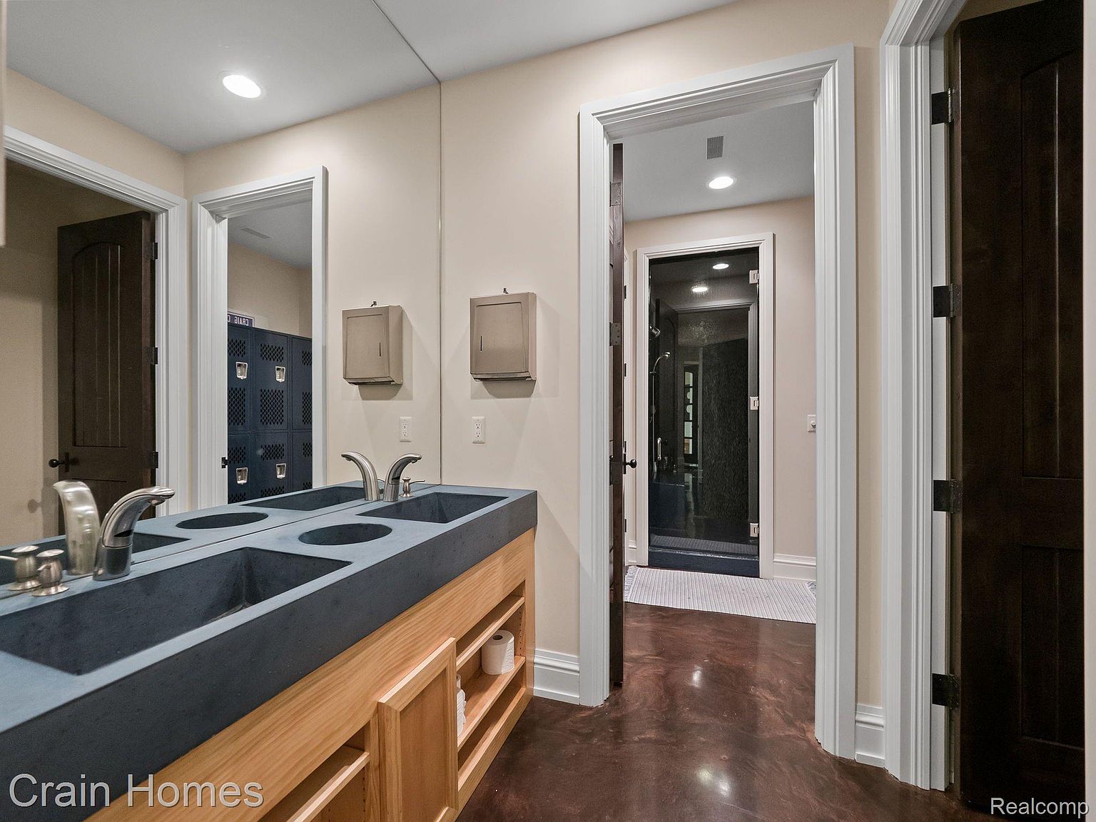 This is a well-lit bathroom featuring a double vanity with a dark countertop and light wood cabinetry. A large mirror reflects the room, showing a glimpse of lockers in an adjacent space. The bathroom also has doorways leading to other areas, including one with a glass-enclosed shower.