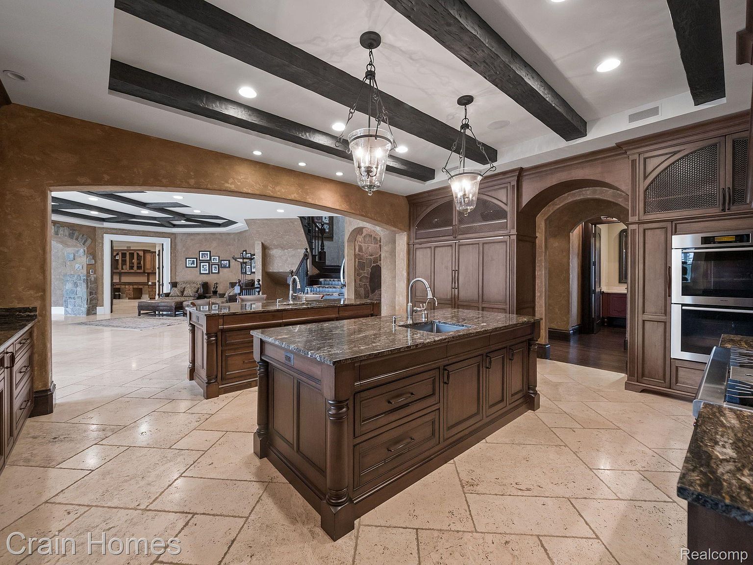 This is a luxurious kitchen featuring two large islands with dark wood cabinetry and granite countertops. The kitchen has dark wood beams on the ceiling and high-end appliances. The flooring is light-colored stone tile, and the kitchen opens to a living area through an arched doorway.