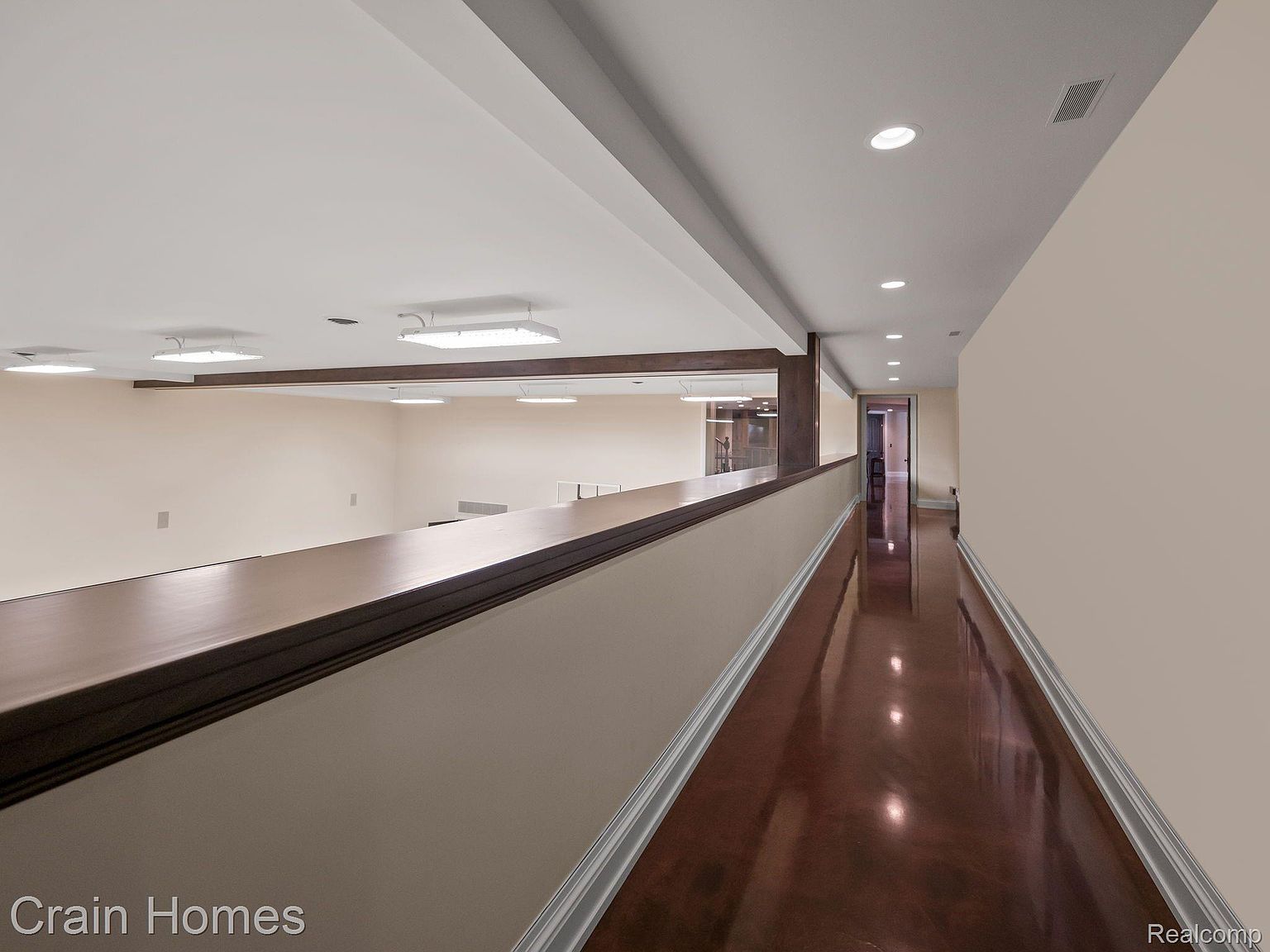 This interior shot showcases a long hallway with a glossy, dark-toned floor reflecting the overhead lighting. The hallway is lined with neutral-colored walls and features a partial view into another room through a framed opening, suggesting a loft or upper level. The overall impression is modern and well-lit, emphasizing the spaciousness of the home.