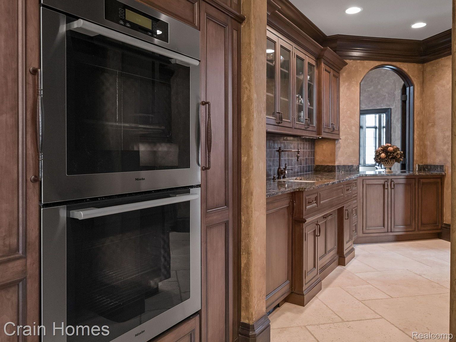 This kitchen features dark wood cabinetry, granite countertops, and stainless steel appliances, including a double oven. The design includes an arched doorway leading to another area, and the flooring is a light-colored tile. The overall impression is one of traditional elegance and functionality.
