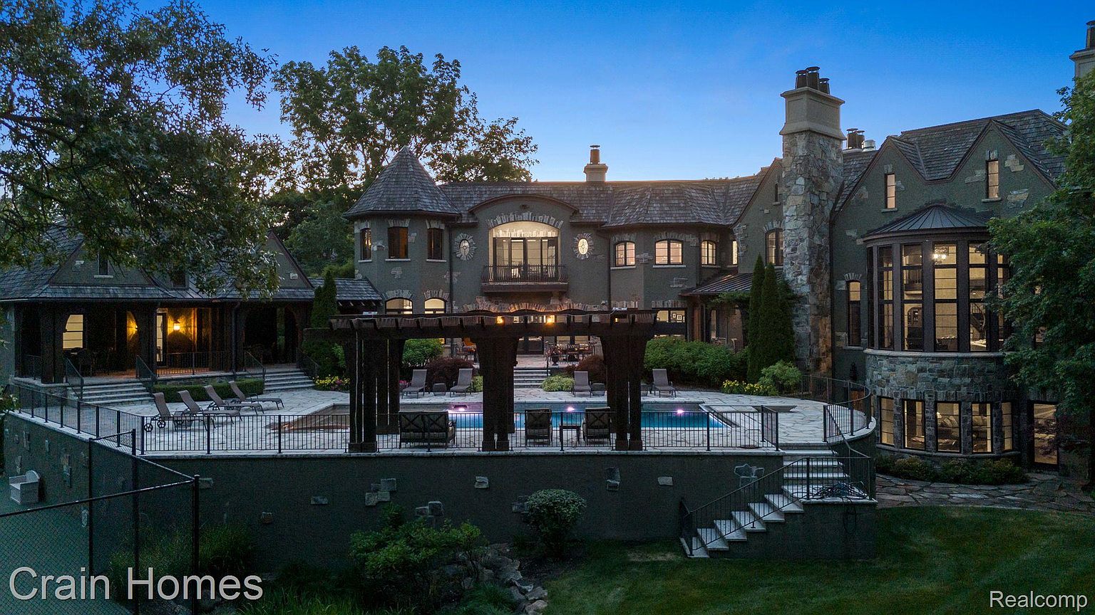 This is a rear exterior view of a large, luxurious home featuring a swimming pool and a meticulously landscaped yard. The architecture includes stone and stucco elements, a turret, and multiple chimneys, creating a stately and elegant impression. A pergola overlooks the pool area, which is surrounded by a patio with lounge chairs.