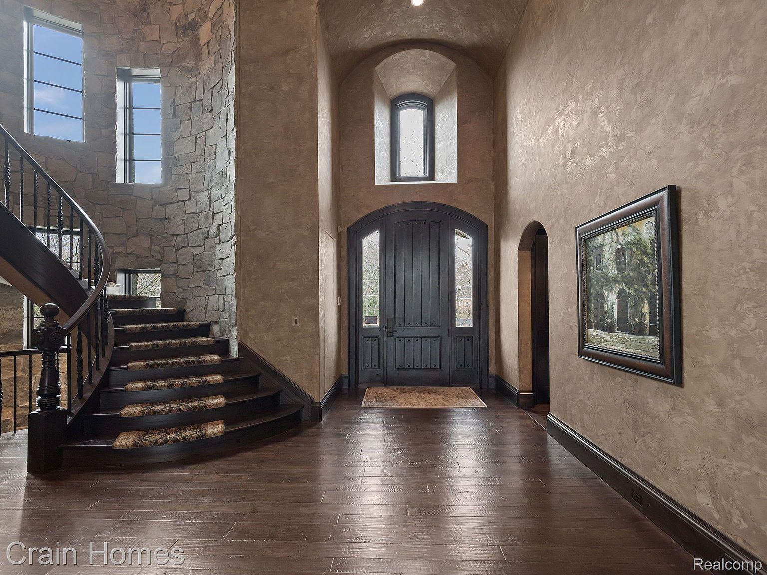 This grand entryway features a curved staircase with carpeted steps and dark wood railings, complemented by a stone wall. The dark wood flooring extends into the hallway, leading to a large wooden front door with an arched window above. A framed painting hangs on the textured wall to the right, adding a touch of elegance.