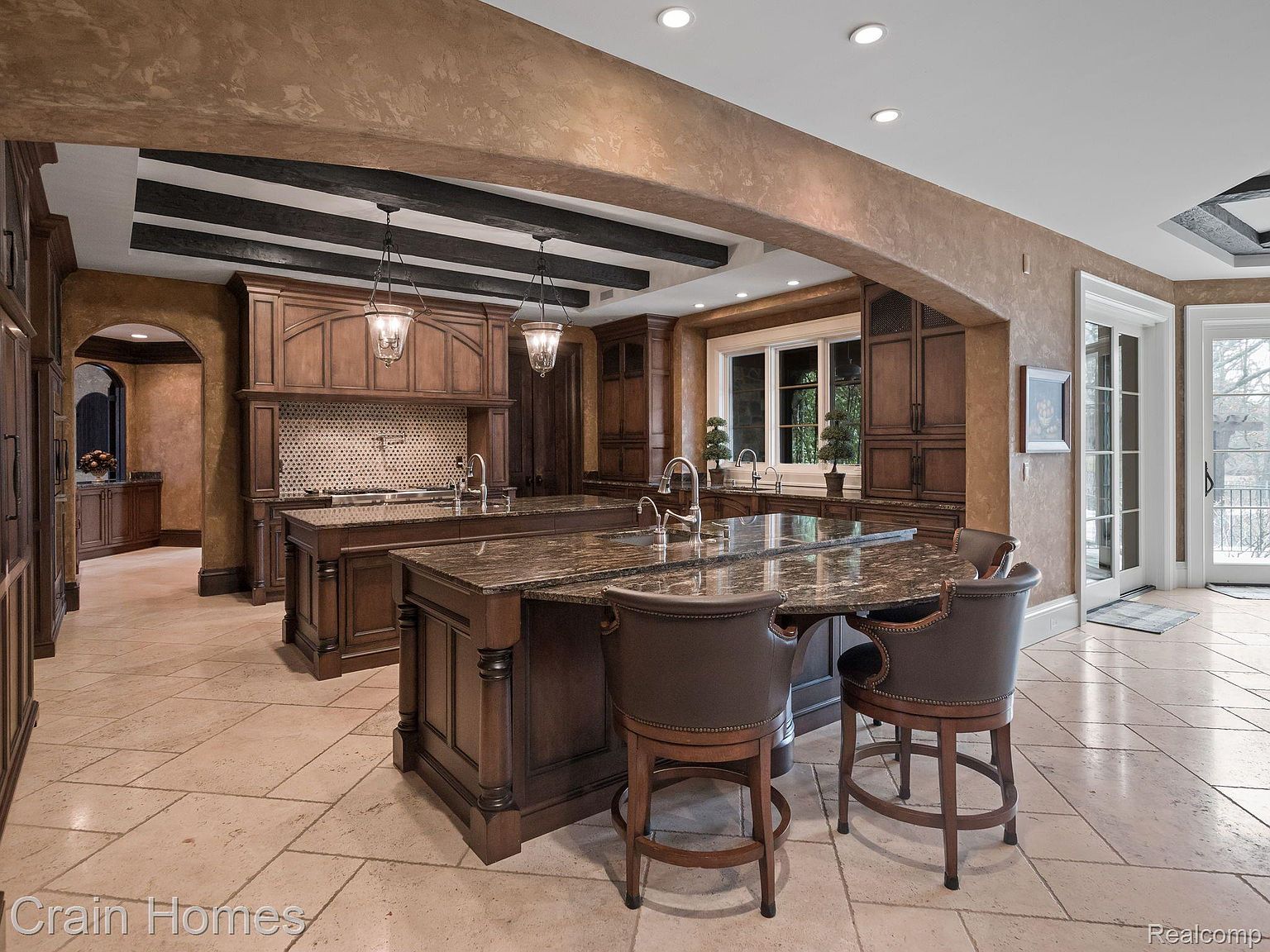 This is a spacious, luxurious kitchen featuring two large islands with granite countertops and dark wood cabinetry. The kitchen is well-lit with recessed lighting and pendant lights, and it has a high-end, traditional style. The flooring is tile, and there is an adjacent dining area visible through an archway.