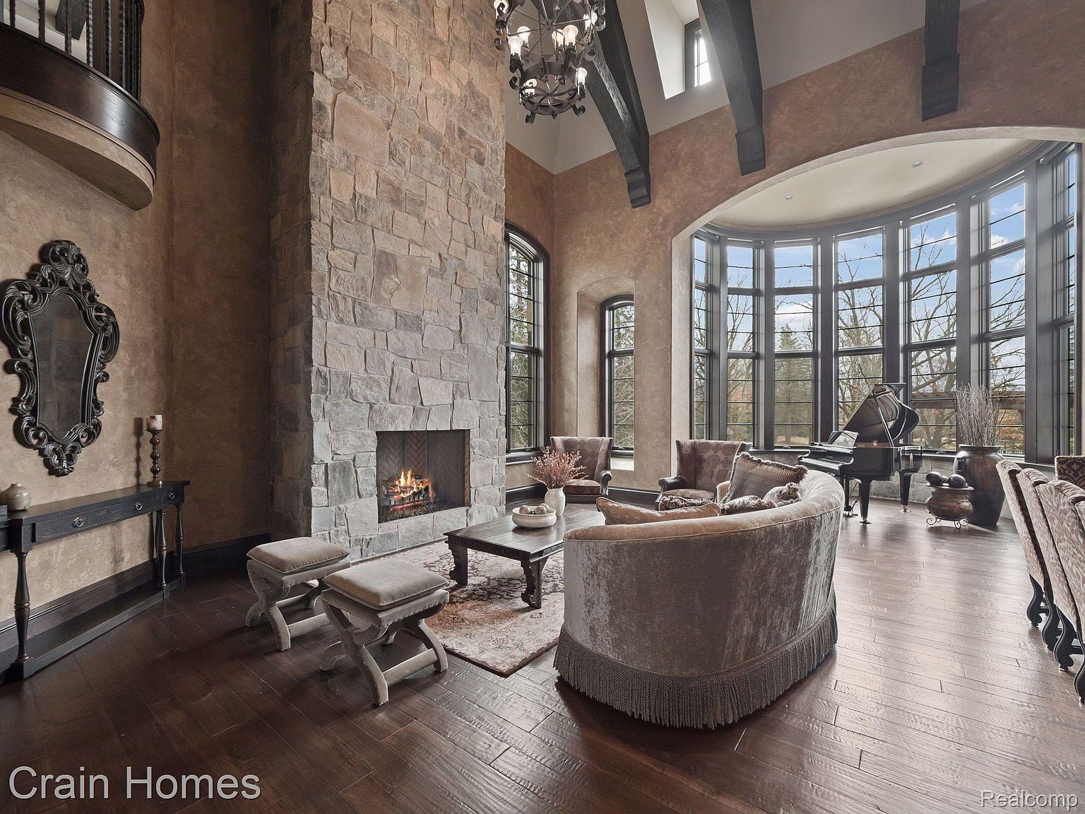This is an interior shot of a luxurious living room featuring a grand stone fireplace as a focal point. The room is furnished with elegant seating, including a curved sofa and armchairs, and a grand piano sits near a large arched window offering natural light. The dark wood flooring and high ceilings with exposed beams contribute to the room's sophisticated and spacious ambiance.