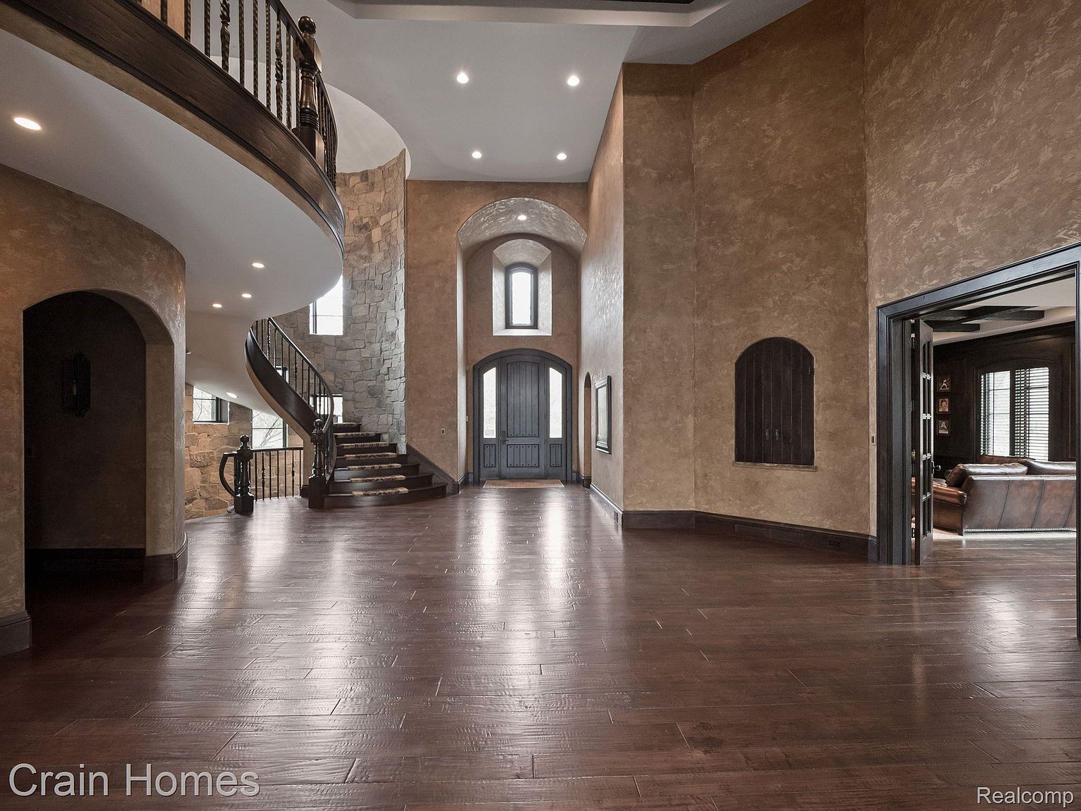 This grand foyer features dark hardwood floors, textured walls, and a curved staircase with wrought iron railings. A stone accent wall adds character, while arched doorways lead to other rooms. The space is well-lit and exudes a luxurious, traditional style.
