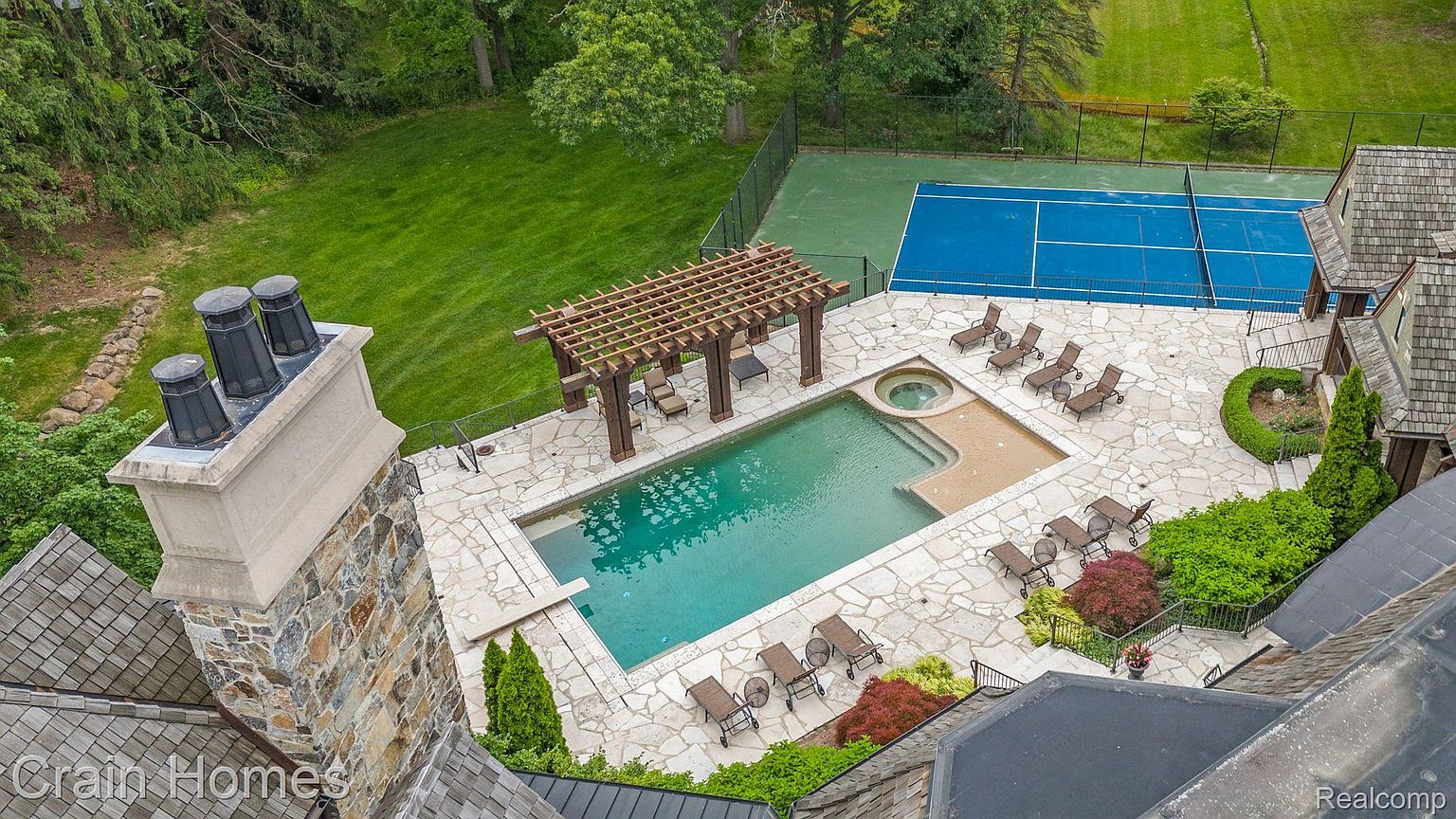 This aerial shot showcases a luxurious backyard featuring a swimming pool, spa, and a pergola-covered lounge area. A tennis court is visible in the background, suggesting ample space for recreation. The stone patio and manicured landscaping add to the property's appeal, creating an inviting outdoor oasis.