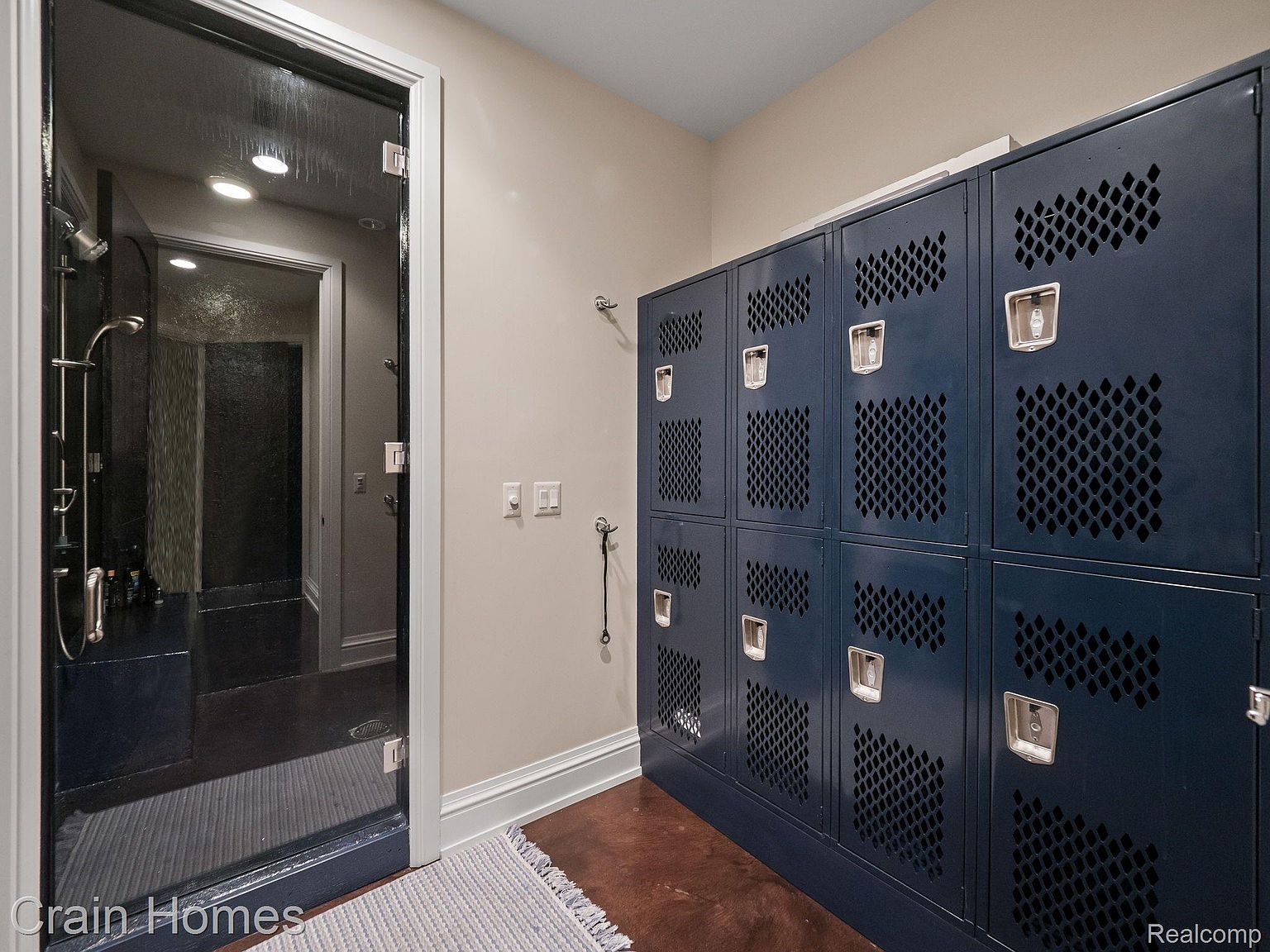 This primary bathroom features a modern shower with dark tile and glass enclosure, complemented by a row of navy blue lockers for storage. The flooring is a rich brown, and a neutral-toned rug adds a touch of comfort. The overall impression is a blend of industrial and contemporary design.