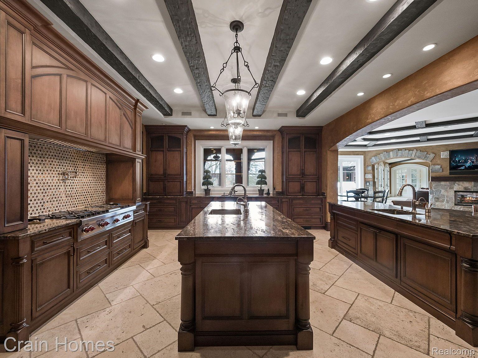 This is a spacious kitchen featuring dark wood cabinetry, granite countertops, and a large central island. The kitchen is well-lit with recessed lighting and a decorative chandelier, and it includes high-end appliances and a tiled backsplash. The overall impression is one of luxury and functionality.