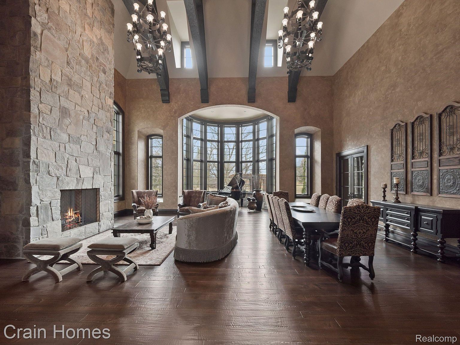 This grand living room features a soaring ceiling with exposed beams and ornate chandeliers, a stone fireplace, and a wall of windows providing ample natural light. The room is furnished with elegant seating arrangements and a long dining table, creating a luxurious and inviting space. The dark wood flooring adds warmth and sophistication to the overall design.