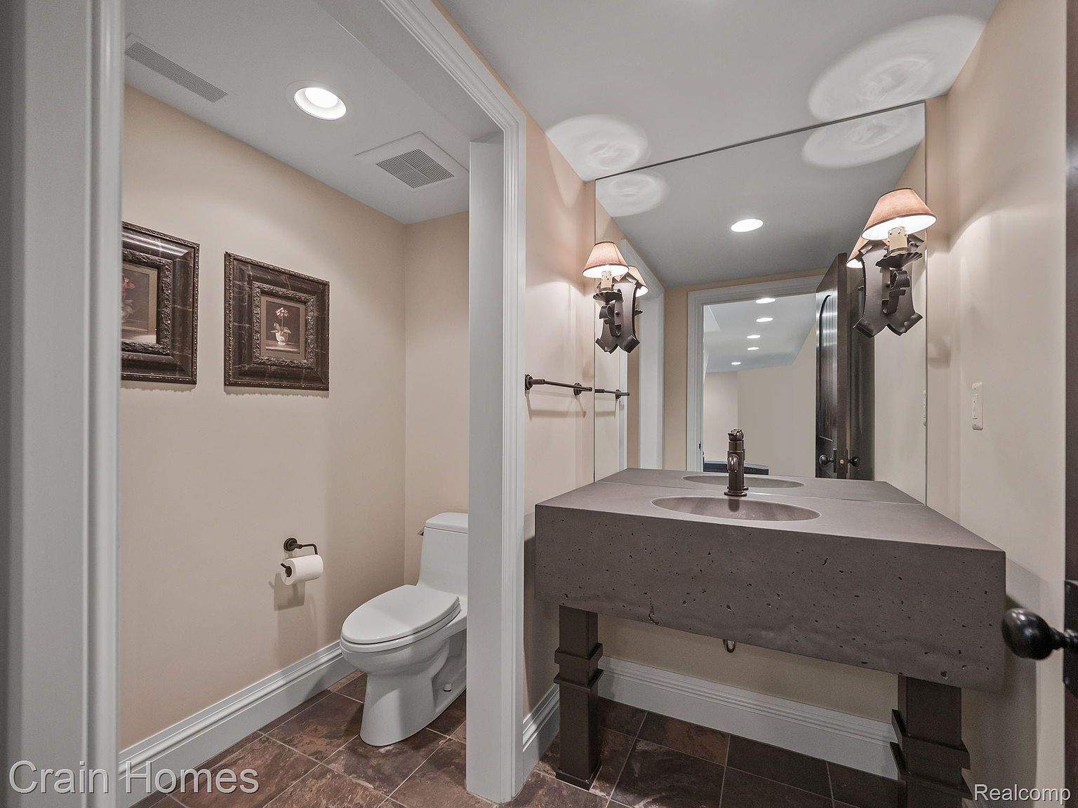 This is a well-appointed guest bathroom featuring a unique concrete double sink vanity with dark wood legs and sconces. The walls are painted in a neutral tone, complemented by dark brown tile flooring. A toilet is visible to the left, and a large mirror reflects the space, enhancing the sense of openness.