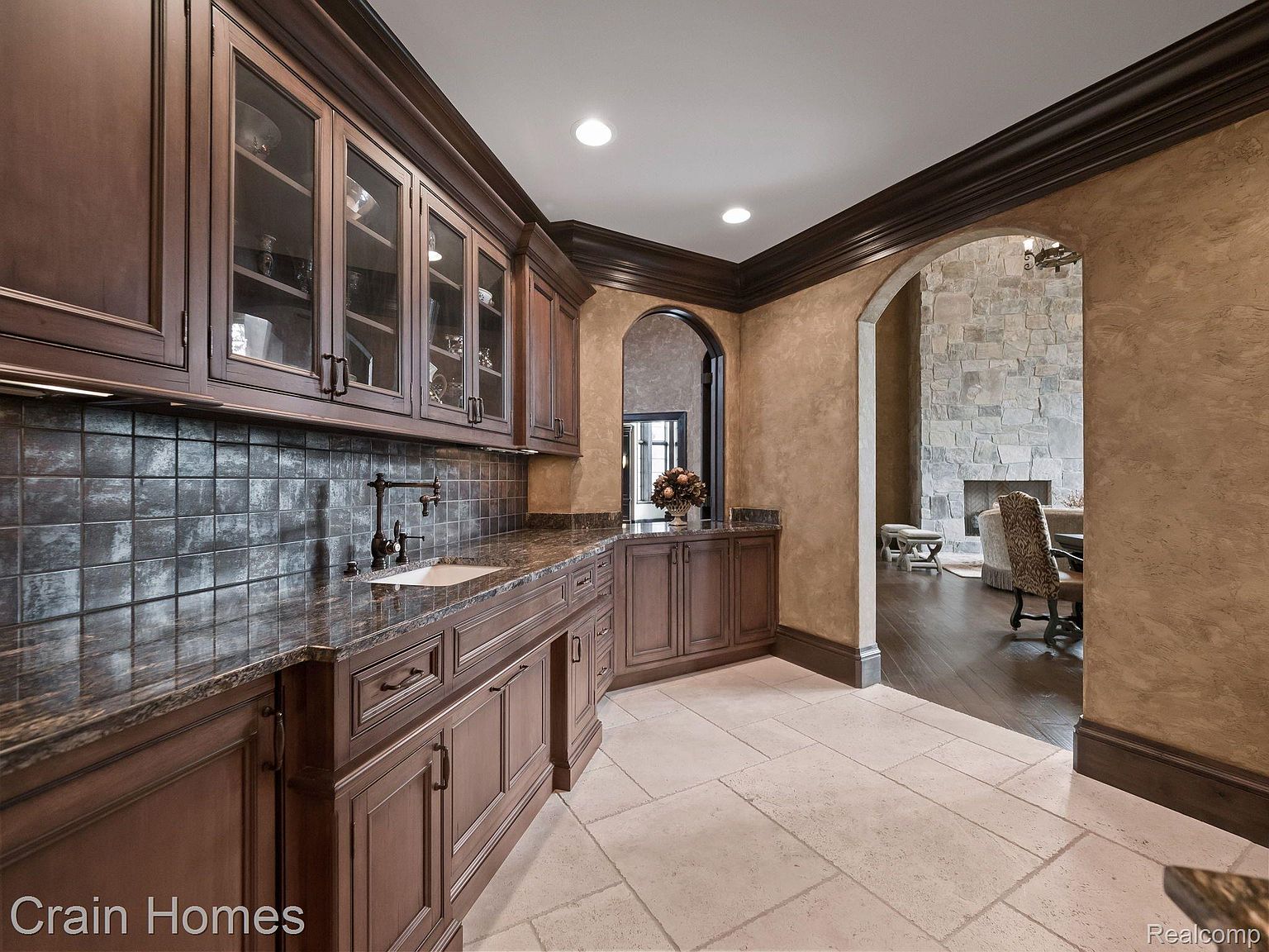 This interior shot showcases a sophisticated kitchen area with dark wood cabinetry, glass-fronted upper cabinets, and a dark countertop. The backsplash features dark, textured tiles, and the flooring is light-colored stone. An arched doorway leads to another room, adding architectural interest and depth to the space.