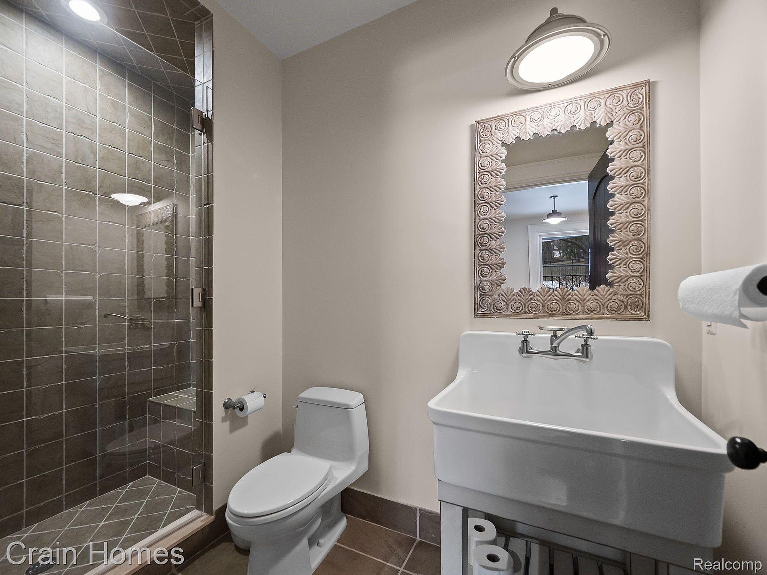 This is a well-lit bathroom featuring a tiled shower with a glass door, a white toilet, and a unique farmhouse-style sink with a decorative mirror above it. The color palette is neutral with brown tile flooring, creating a clean and functional space. The perspective is from the doorway, showcasing the entire bathroom layout.