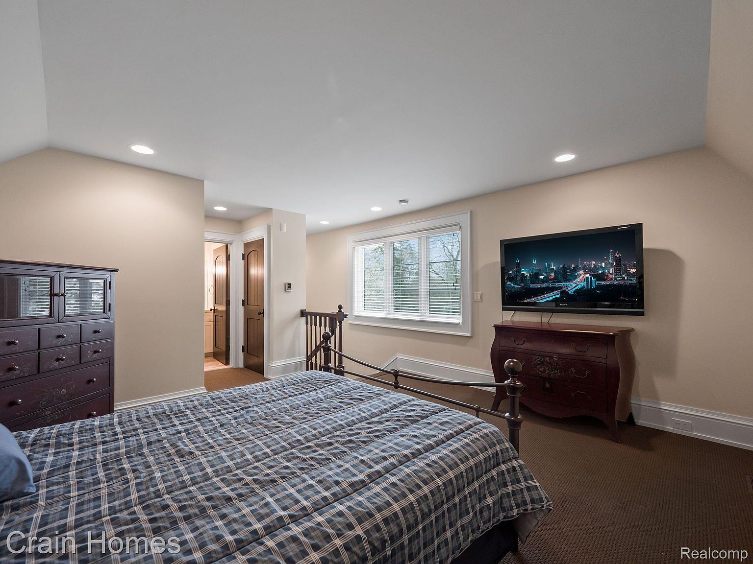 This is a cozy bedroom featuring a plaid-covered bed, a dark wood dresser, and a flat-screen TV mounted on the wall. A window with blinds provides natural light, and the room has neutral-colored walls and carpet. The overall impression is comfortable and inviting.