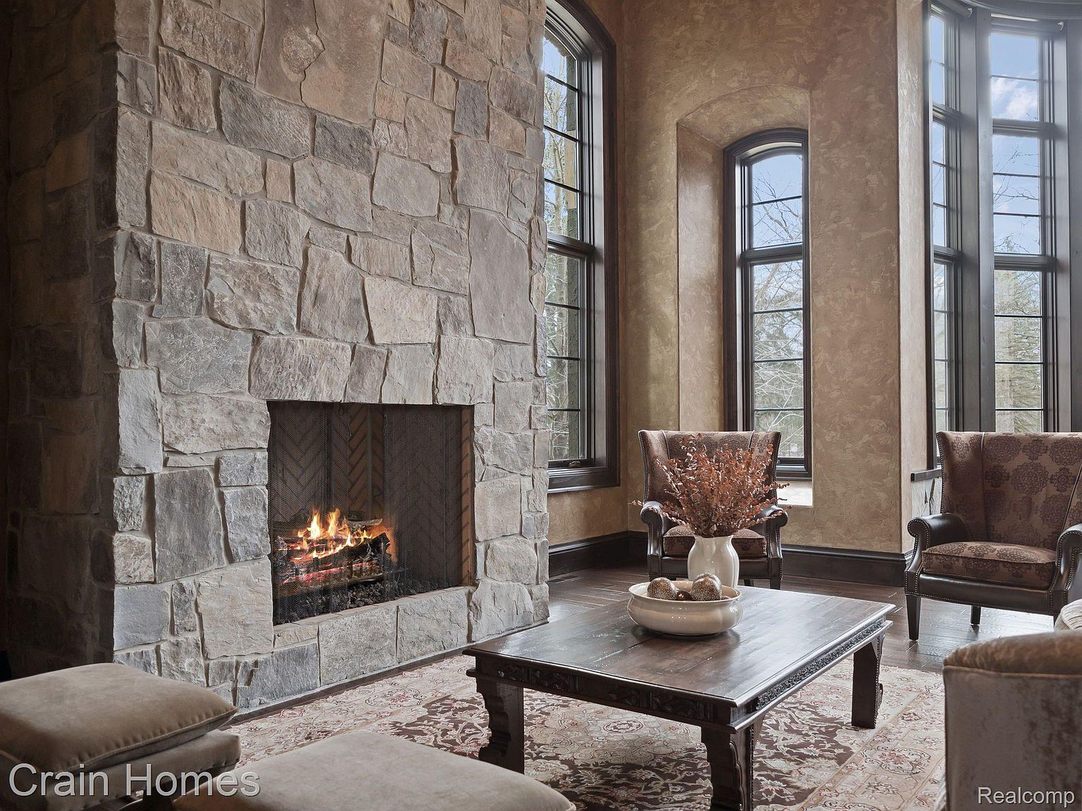 This interior shot showcases a luxurious living room with a grand stone fireplace as the focal point. The room features elegant armchairs, a dark wood coffee table with decorative elements, and large windows that provide ample natural light. The overall impression is one of warmth, sophistication, and classic design.