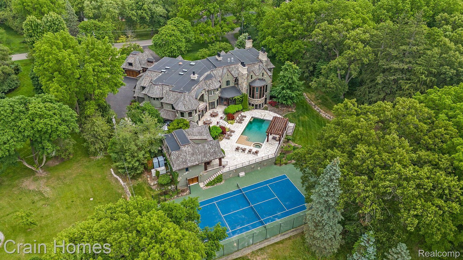 This aerial shot showcases a sprawling estate with a unique architectural design, featuring a combination of stone and shingle roofing. The property includes a swimming pool, tennis court, and meticulously landscaped grounds with mature trees providing privacy. The overall impression is one of luxury and seclusion.