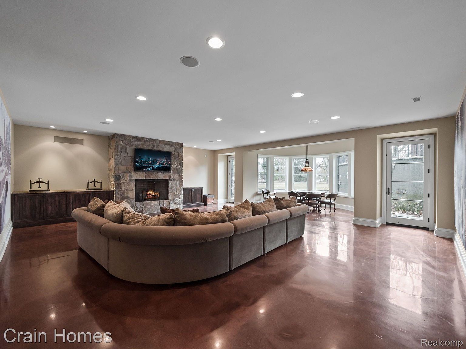 This is a spacious living room featuring a large, curved sofa facing a stone fireplace with a mounted television. The room has a polished, reflective floor and recessed lighting, creating a bright and open atmosphere. A dining area is visible in the background, adding to the sense of expansive living space.