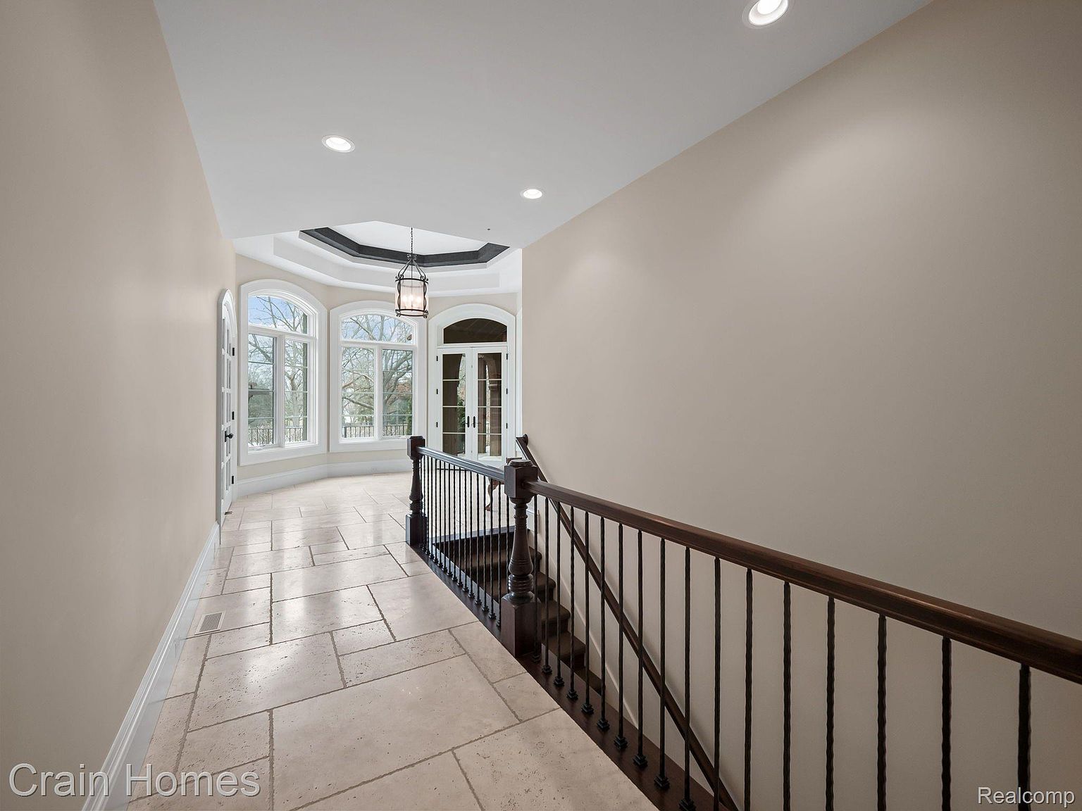 This interior shot showcases a hallway with stairs, featuring light beige walls and tiled flooring. Large arched windows at the end of the hallway provide natural light and a view to the outside. A dark wood railing and wrought iron balusters line the staircase, adding a touch of elegance.