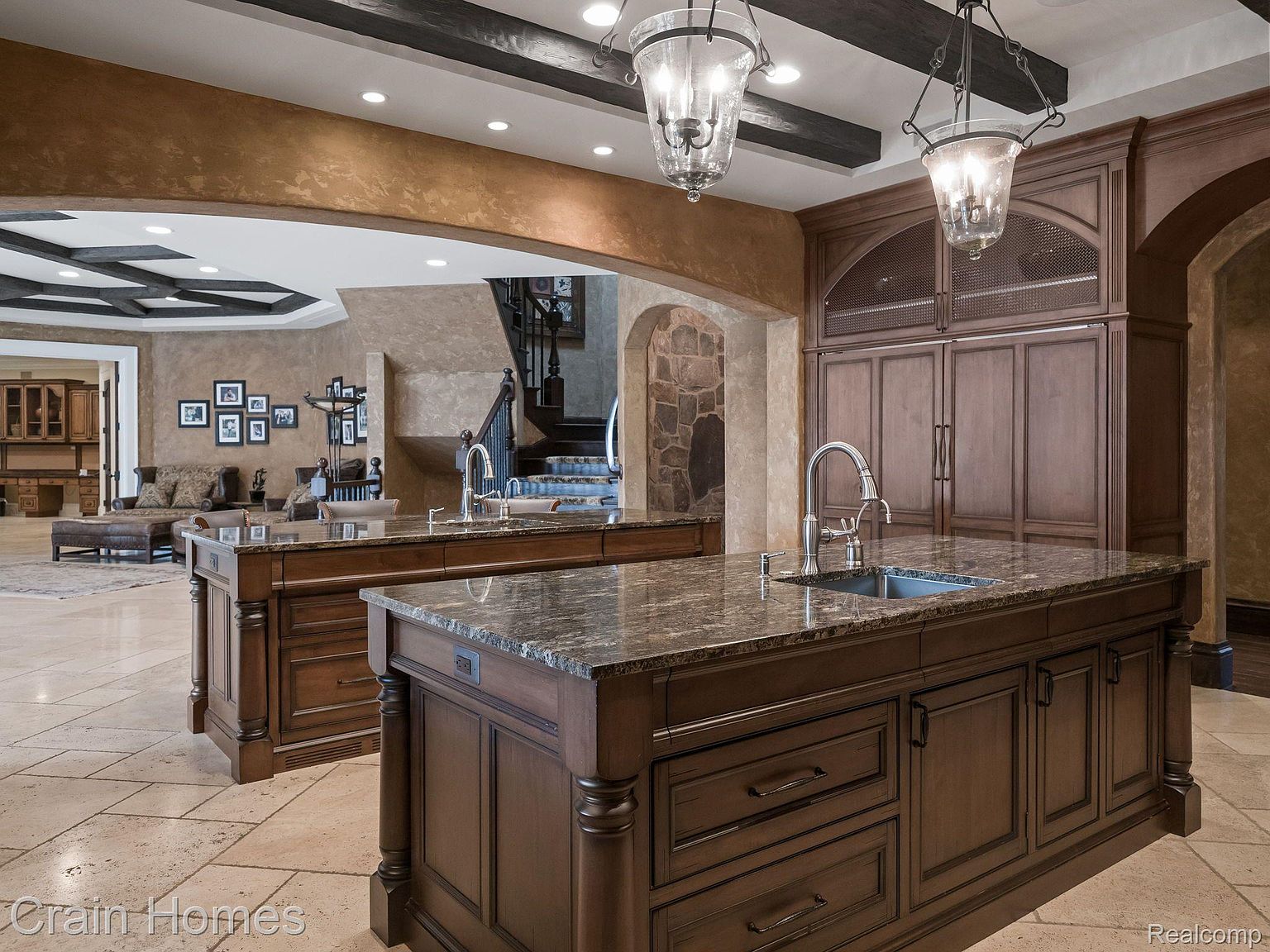 This interior shot showcases a luxurious kitchen with two large islands featuring dark wood cabinetry and granite countertops. Ornate pendant lights hang above the islands, and the kitchen seamlessly transitions into a living area with a staircase visible in the background. The overall impression is one of spaciousness and high-end design.