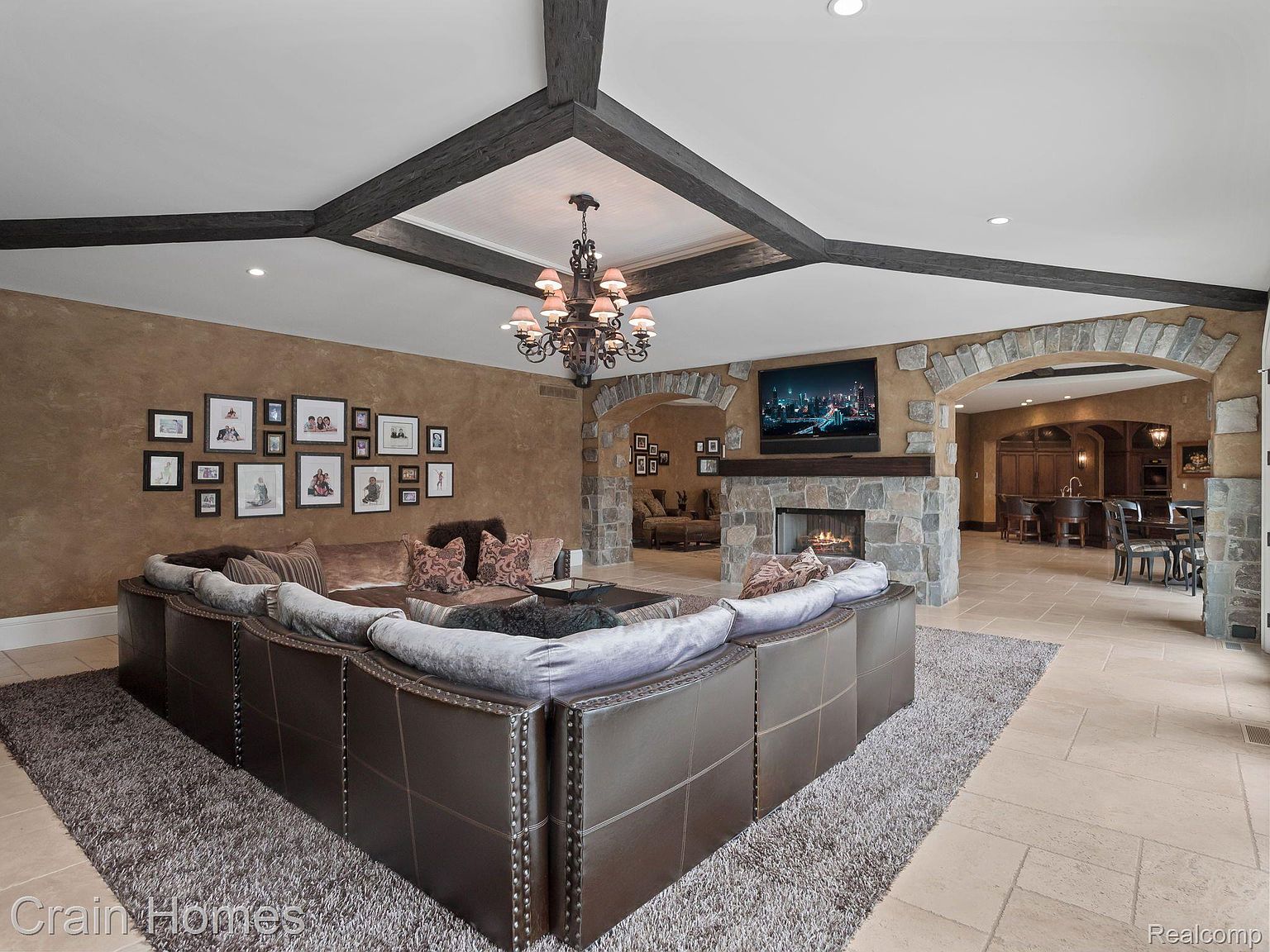 This is an interior shot of a luxurious living room featuring a large, brown leather sectional sofa with studded detailing, set on a gray shag rug. The room has a stone fireplace with a mounted television above it, and a stone archway leads to another room with a bar area. The ceiling has dark wooden beams and a decorative chandelier, creating a warm and inviting atmosphere.