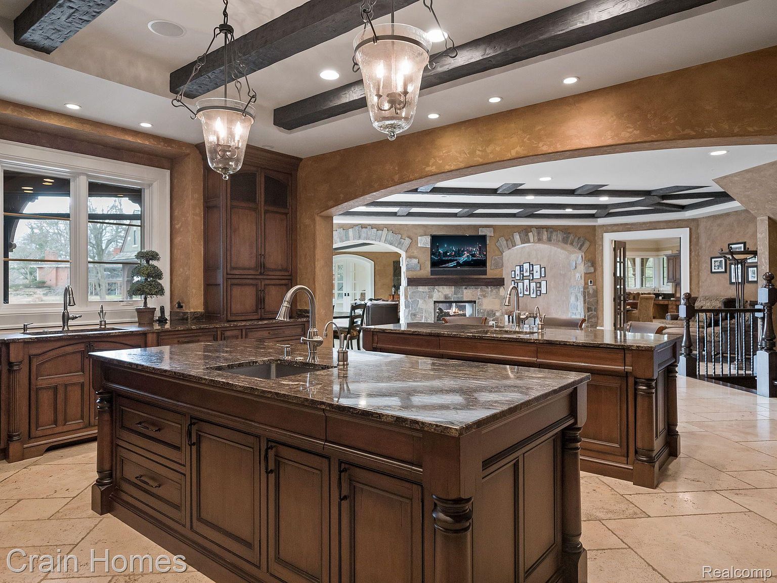 This is a luxurious kitchen featuring two large islands with granite countertops and dark wood cabinetry. The kitchen has high-end appliances, decorative pendant lighting, and an open layout that connects to other living spaces through an arched entryway. The overall impression is one of elegance and spaciousness, ideal for entertaining.