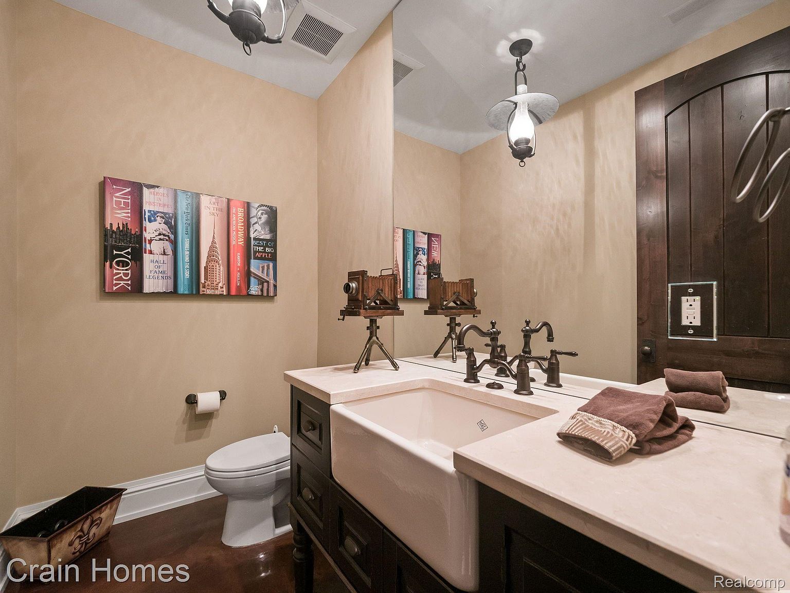 This is a well-appointed guest bathroom featuring a unique design. The walls are painted in a warm, neutral tone, complemented by dark wood accents on the vanity and door. A farmhouse-style sink sits atop a light-colored countertop, and vintage-inspired fixtures add character. A decorative book display and antique camera add a touch of personality.