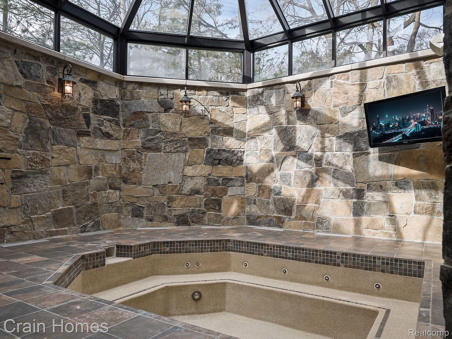 This sunroom features a large jacuzzi tub surrounded by tile and stone walls, creating a luxurious spa-like atmosphere. The room is enclosed by a glass ceiling and windows, providing ample natural light and views of the surrounding trees. A wall-mounted television adds a modern touch to this relaxing space.
