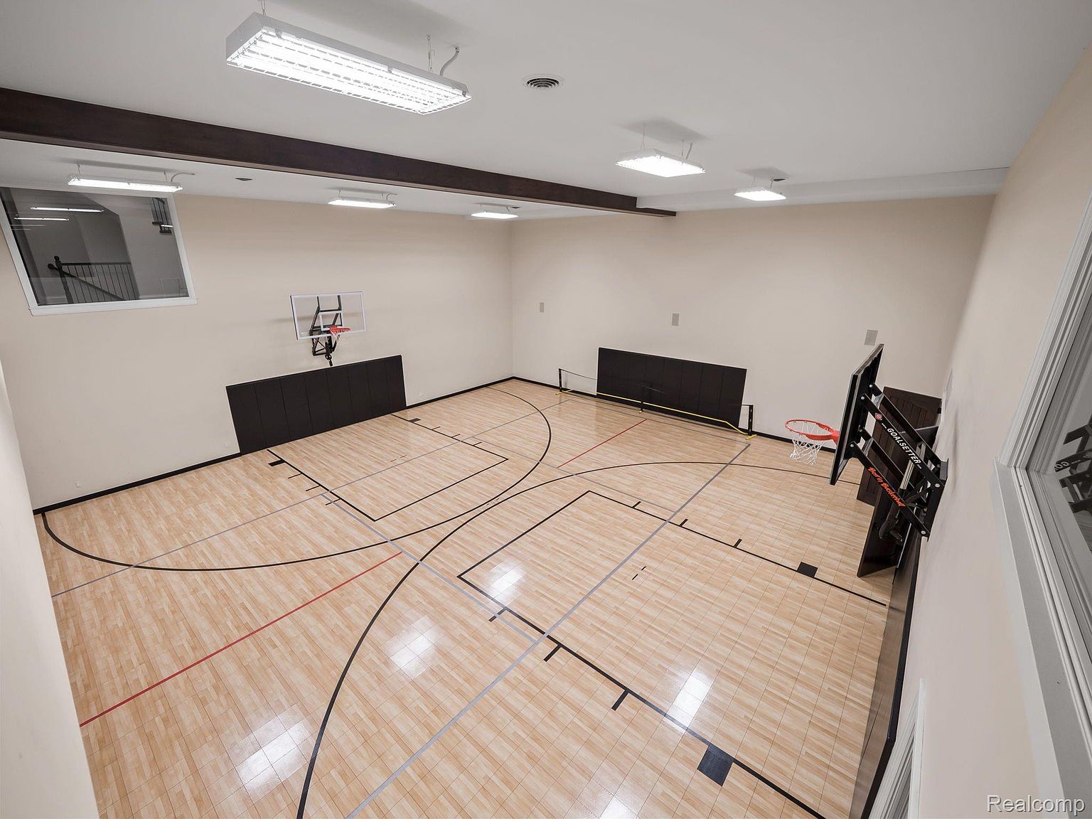 This is an interior shot of a home gym featuring a basketball court. The court has a light-colored wood floor with black lines marking the boundaries and key areas. There are two basketball hoops, one mounted on the wall and another portable one, and the walls are painted in a neutral tone, with dark padding along the lower sections.