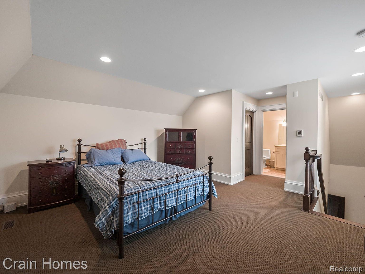 This is a cozy bedroom featuring a wrought iron bed with blue bedding and a plaid comforter. The room has a warm, inviting feel with neutral-colored walls and brown carpeting. A dark wood dresser and chest of drawers provide ample storage, and a doorway leads to an adjacent bathroom, suggesting a private suite.