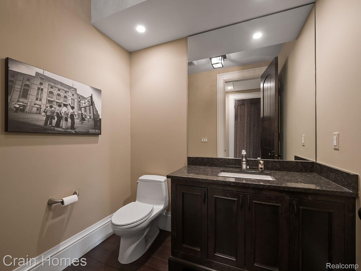 This is a cozy guest bathroom featuring a toilet, a dark wood vanity with a granite countertop, and a large mirror reflecting a doorway. The walls are painted in a warm beige tone, and a black and white photograph hangs above the toilet, adding a touch of character. The overall impression is clean and functional, with a classic design.