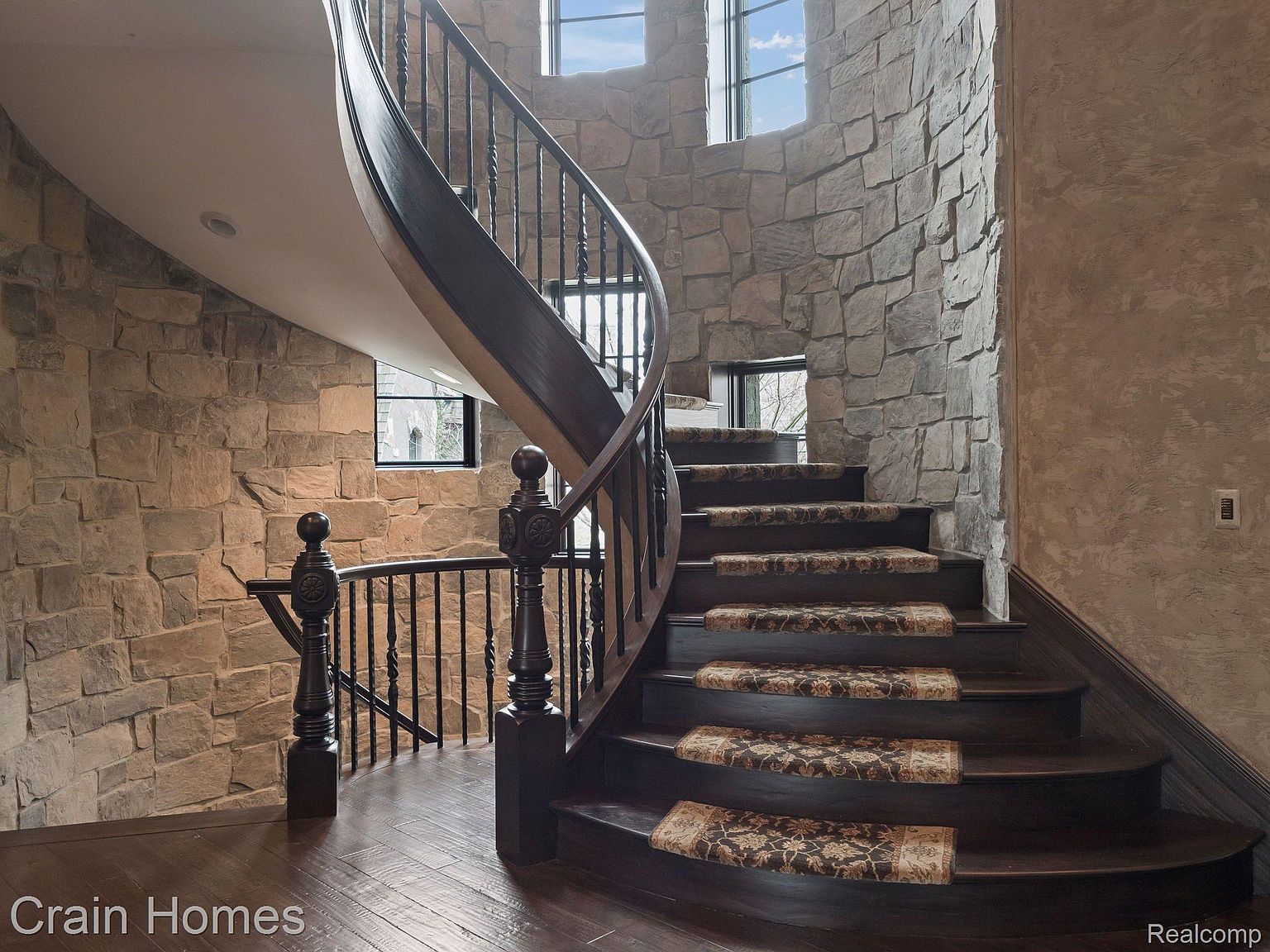 This interior shot showcases a grand, curved staircase with dark wood steps and ornate railings. The walls are a mix of stone and textured plaster, creating a rustic yet elegant ambiance. Natural light streams in through strategically placed windows, highlighting the architectural details and the craftsmanship of the staircase.